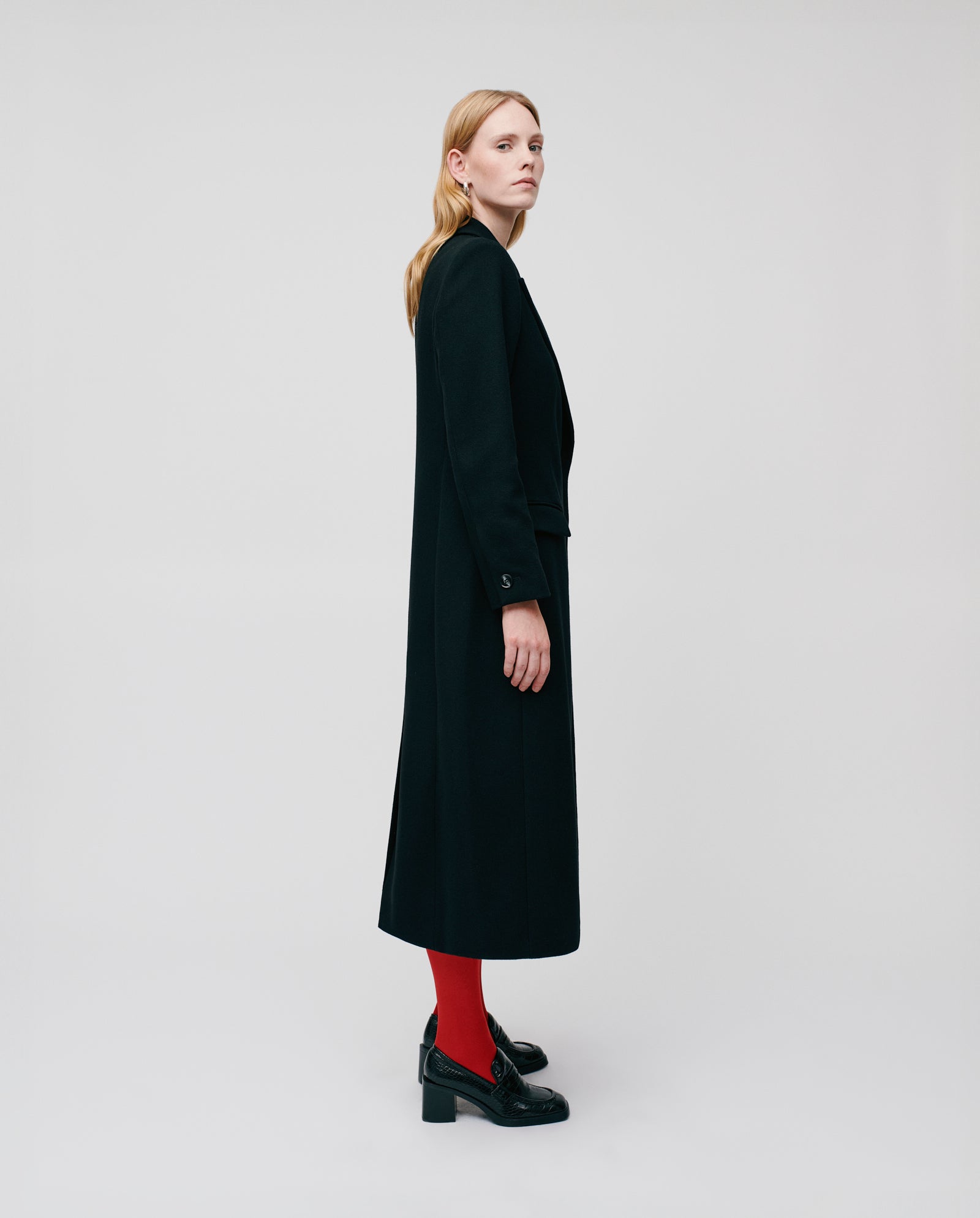 A woman stands sideways against a plain white background wearing the IVY OAK CYNTHIA Coat, a long black luxury women’s coat with a fitted waist, paired with red tights.