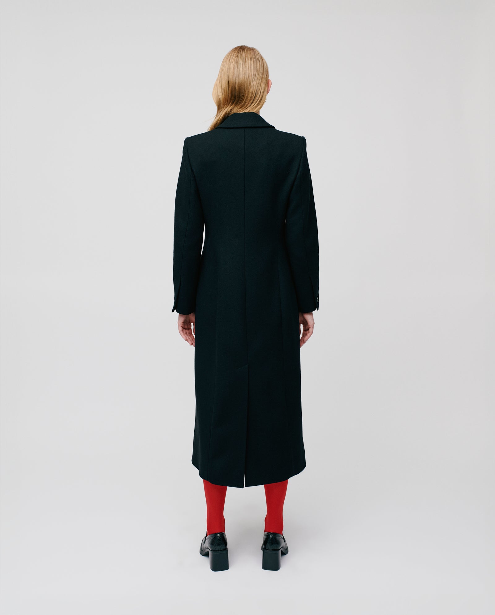 A person wearing the IVY OAK CYNTHIA Coat—a luxury women's virgin wool coat with a fitted waist—paired with red tights and black shoes stands facing away against a plain background.