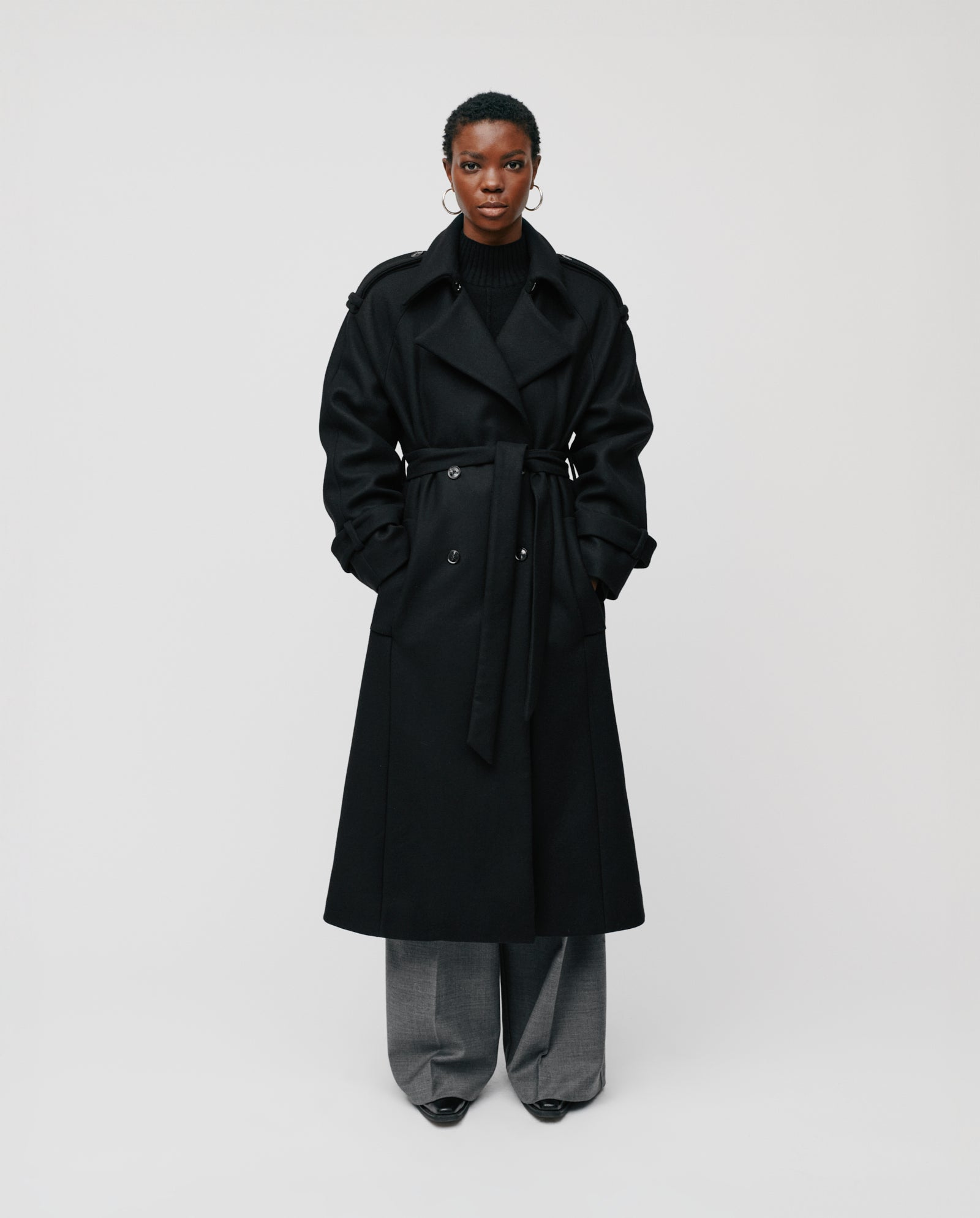 A person in the IVY OAK CAROLINE ANN long black winter coat crafted from RWS-certified wool, layered over gray wide-leg pants, stands against a plain white background.