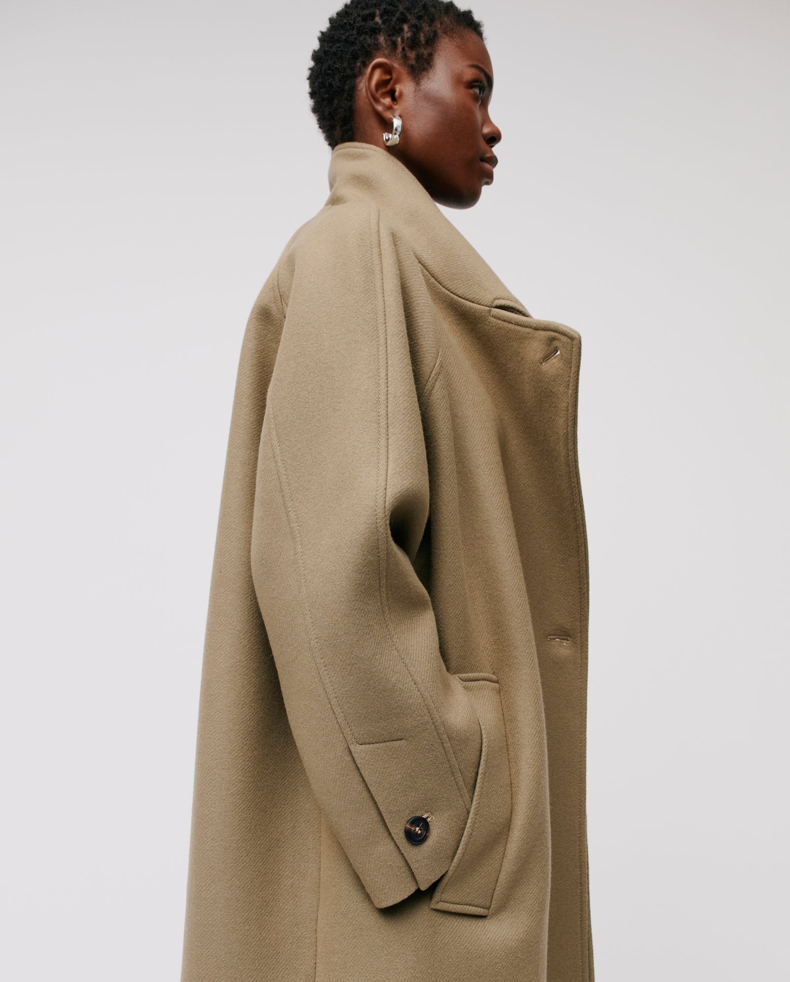 A person wears the IVY OAK CLEO Coat, featuring an oversized fit, high stand-up collar, and raglan sleeves, looking to the side against a plain background.