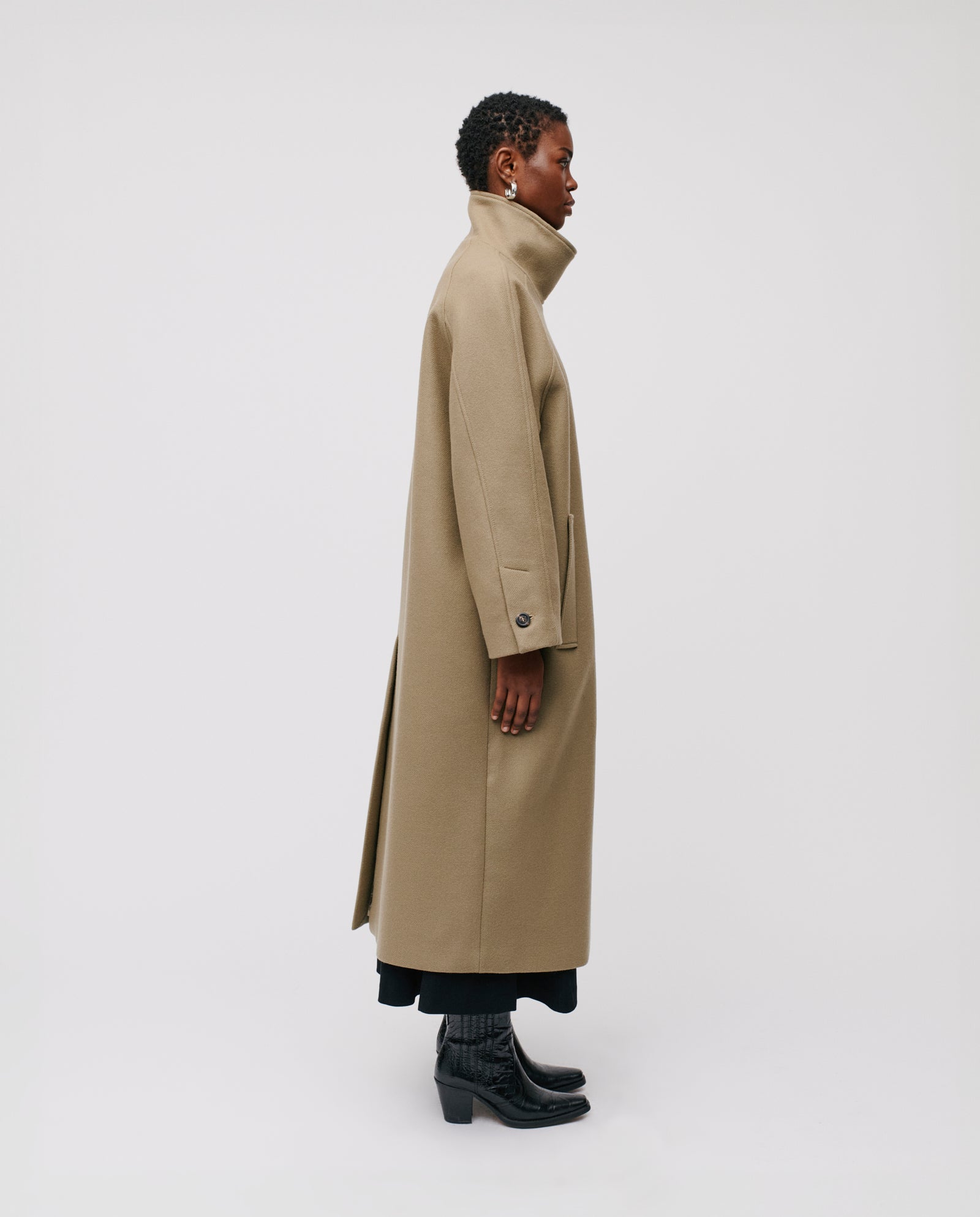 A person stands in profile against a plain white background wearing the IVY OAK CLEO Coat—an oversized tan caban style with a high stand-up collar—paired with black pants and boots.