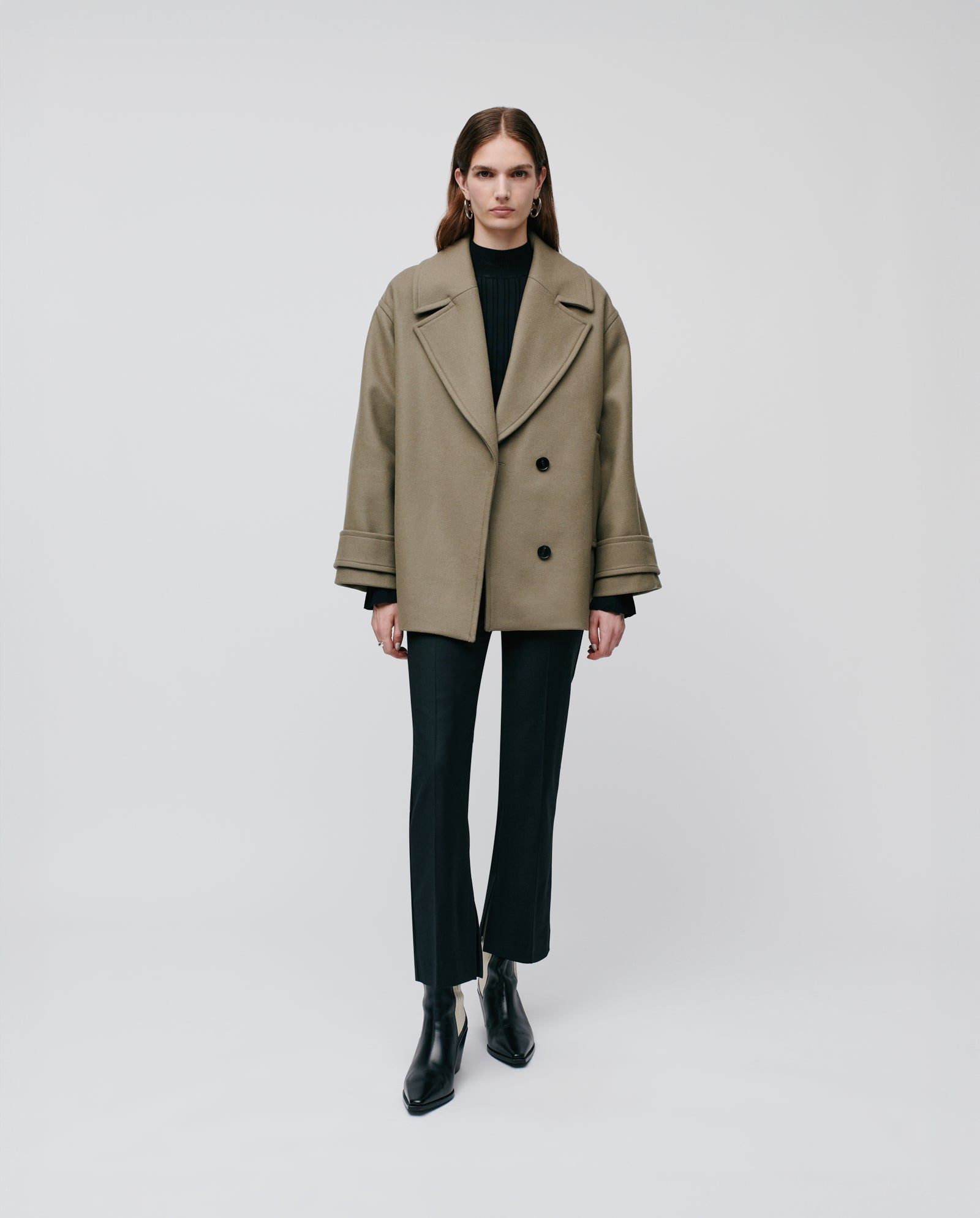 A woman models the IVY OAK CARLY Pea Coat, an olive oversized jacket with hidden button fastening, styled with black pants and ankle boots against a plain backdrop.