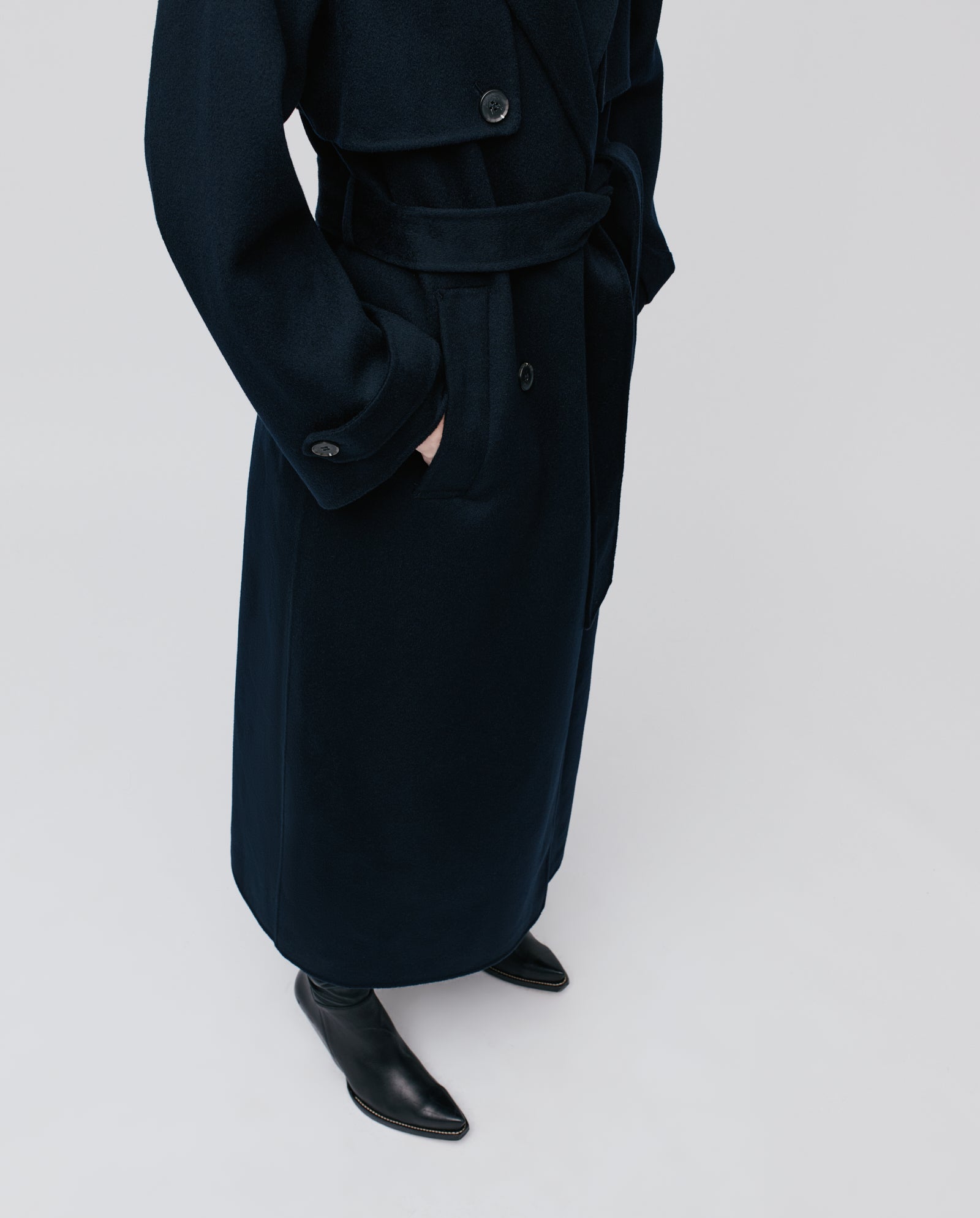 A person wearing the IVY OAK COLETTE ANN oversized dark trenchcoat made from Responsible Wool and black pointed boots stands with hands in pockets against a plain light background.
