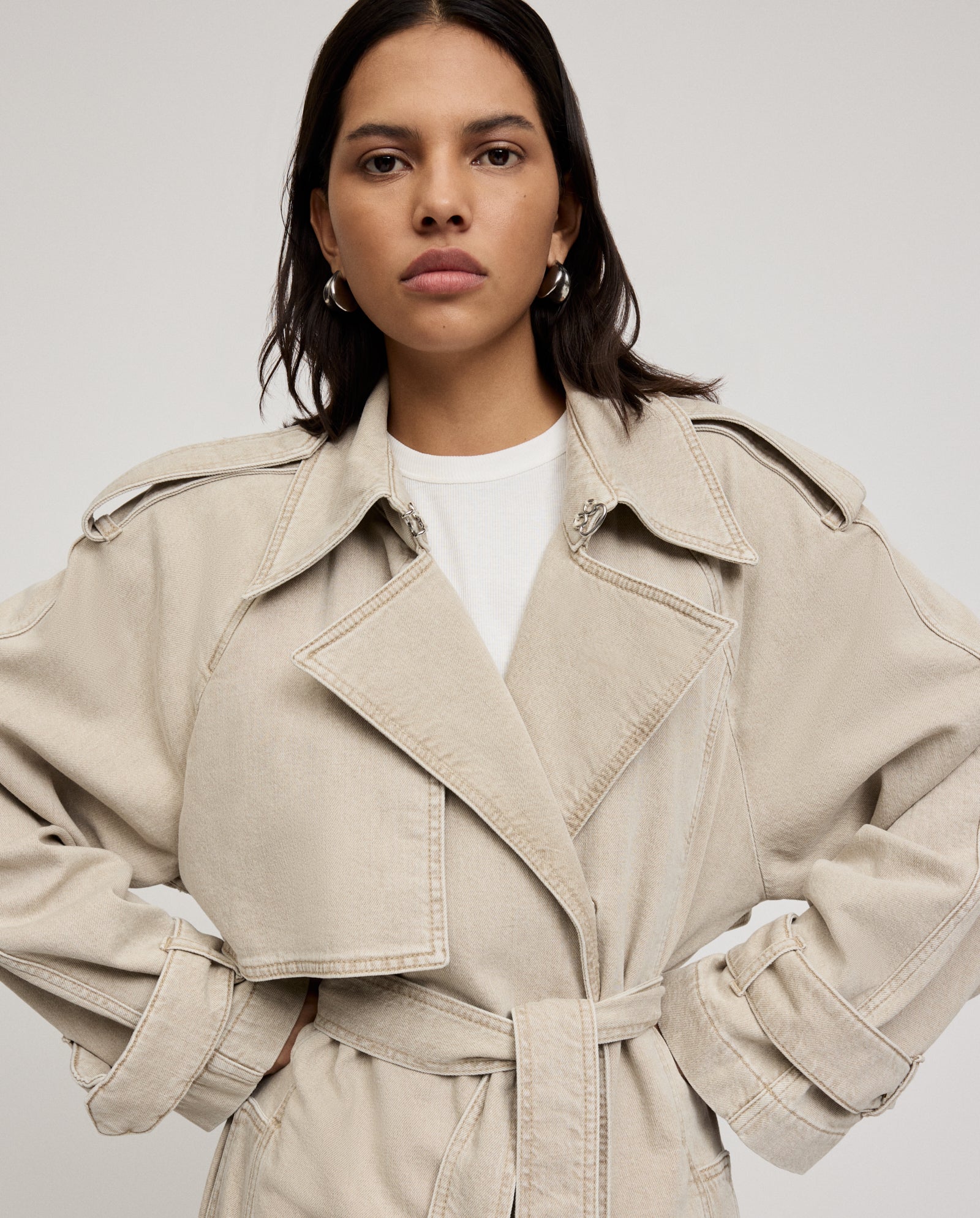 A woman stands confidently with hands on hips, wearing the IVY OAK CORDELIA Coat—an oversized beige trench with front pockets and an adjustable belt—over a white shirt.