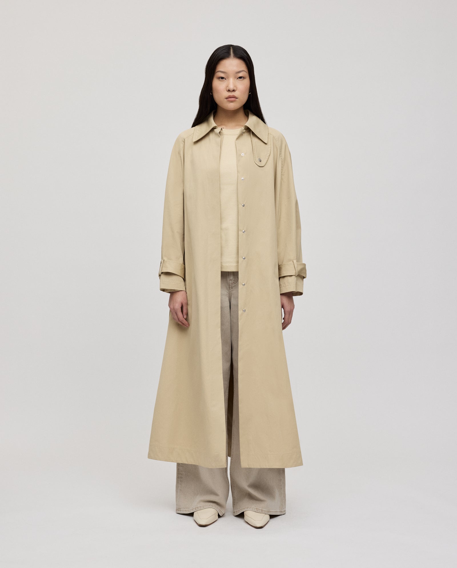 A woman wears the IVY OAK CLEMENTINE Coat, a long beige A-line trench with silver accents, paired with wide-leg pants and light shoes, standing against a plain background.