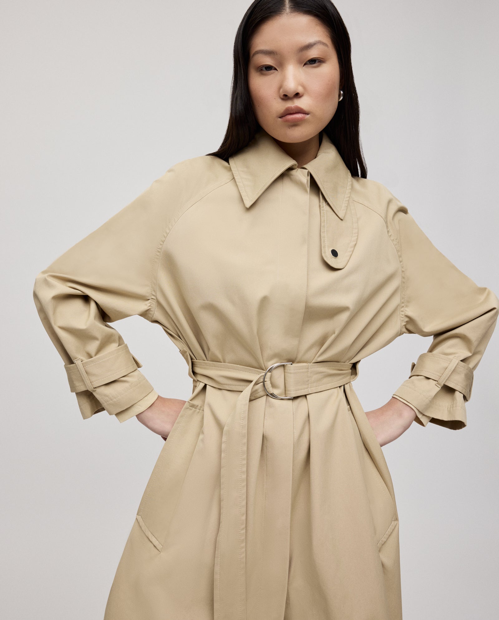 A woman poses hands on hips in the IVY OAK CLEMENTINE A-line trench coat with silver accents, set against a plain background.