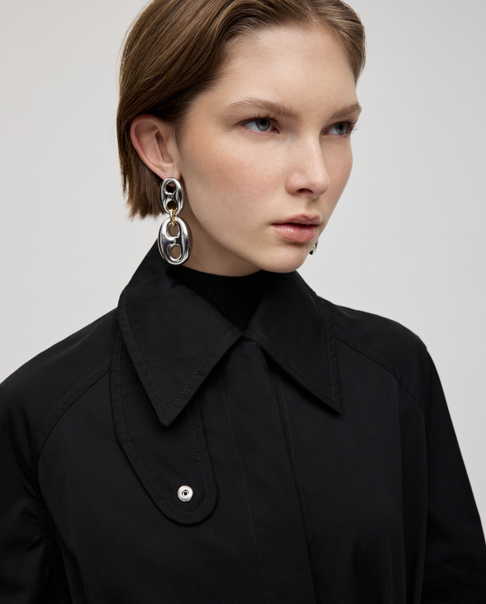A woman with short light brown hair wears the IVY OAK CLEMENTINE Coat, a black A-line trench with silver accents, styled with large silver statement earrings.