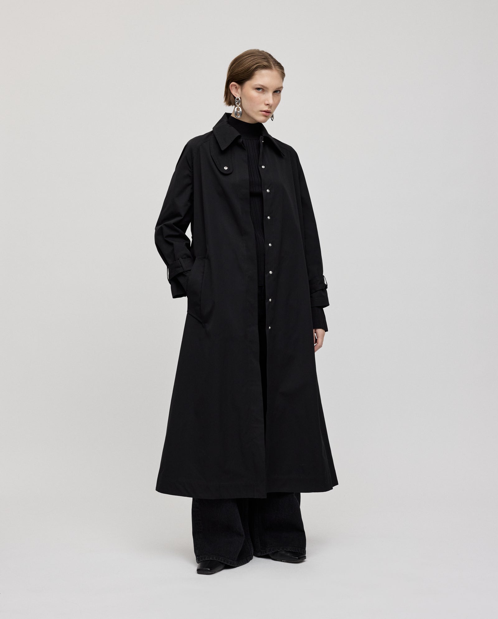 A person wears the IVY OAK CLEMENTINE Coat, a long black trench with subtle silver accents, over black pants against a plain light background.