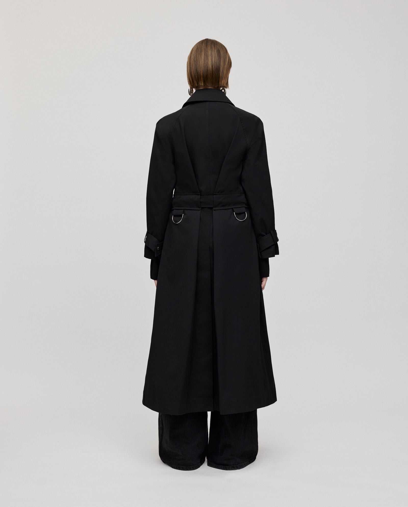 A person faces away, wearing IVY OAKs CLEMENTINE A-line trench coat with black pants against a plain light background.