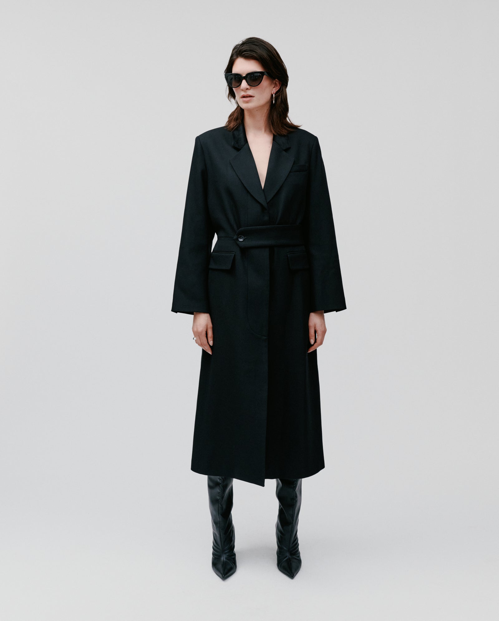 A woman stands against a plain background wearing the IVY OAK CORI Coat, paired with sunglasses and black knee-high boots.