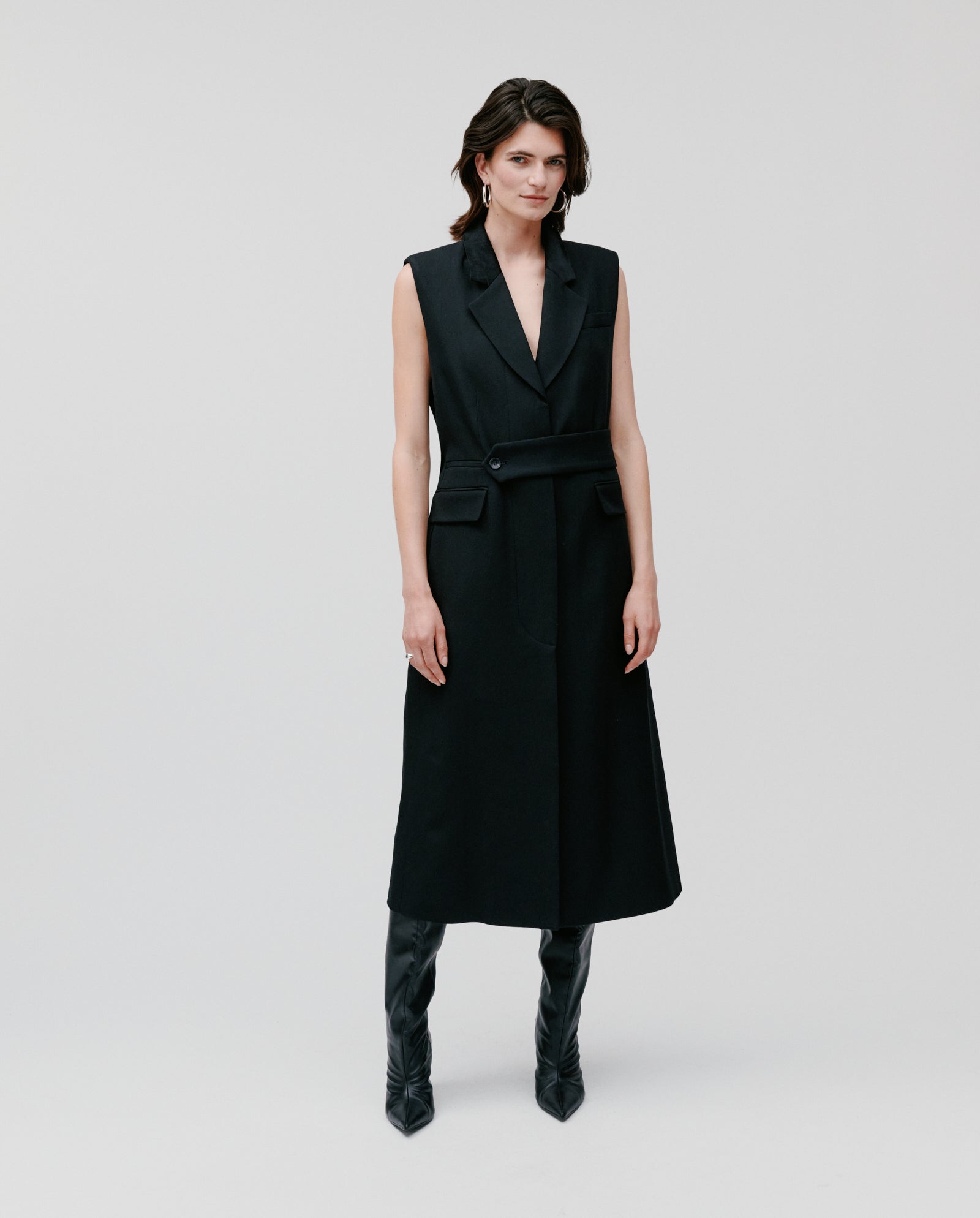 A woman wearing the IVY OAK CORI ANN Vest, a sleeveless black belted dress with an adjustable belt, paired with tall black boots, stands against a plain light background.