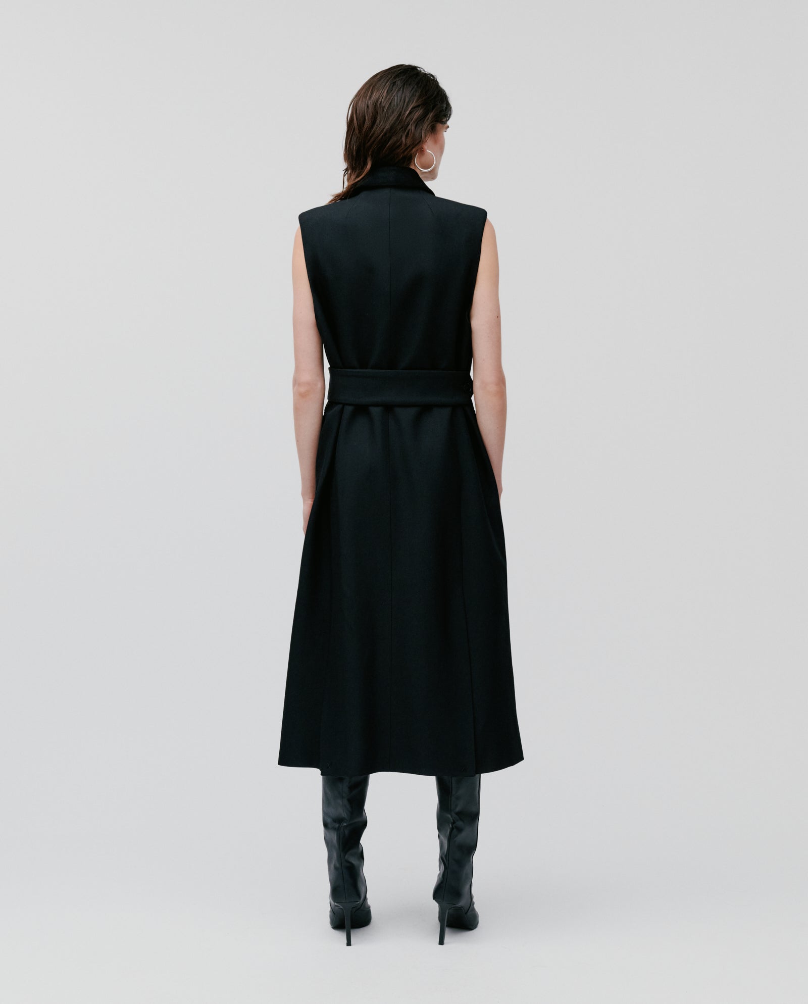 A person seen from the back wears the IVY OAK CORI ANN Vest, a sleeveless black trench coat with an adjustable belt, paired with tall black boots against a plain background.