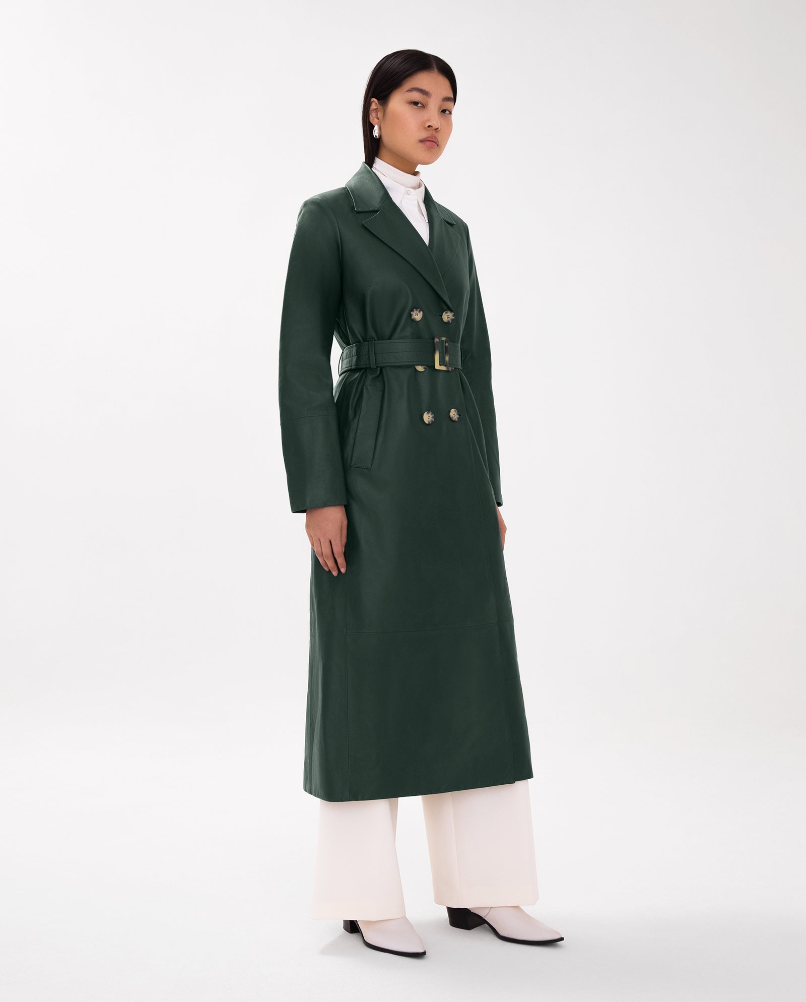 A woman stands against a plain background wearing the IVY OAK LIONNE Coat, a long dark green leather trench in midi length, styled over white pants and shoes.