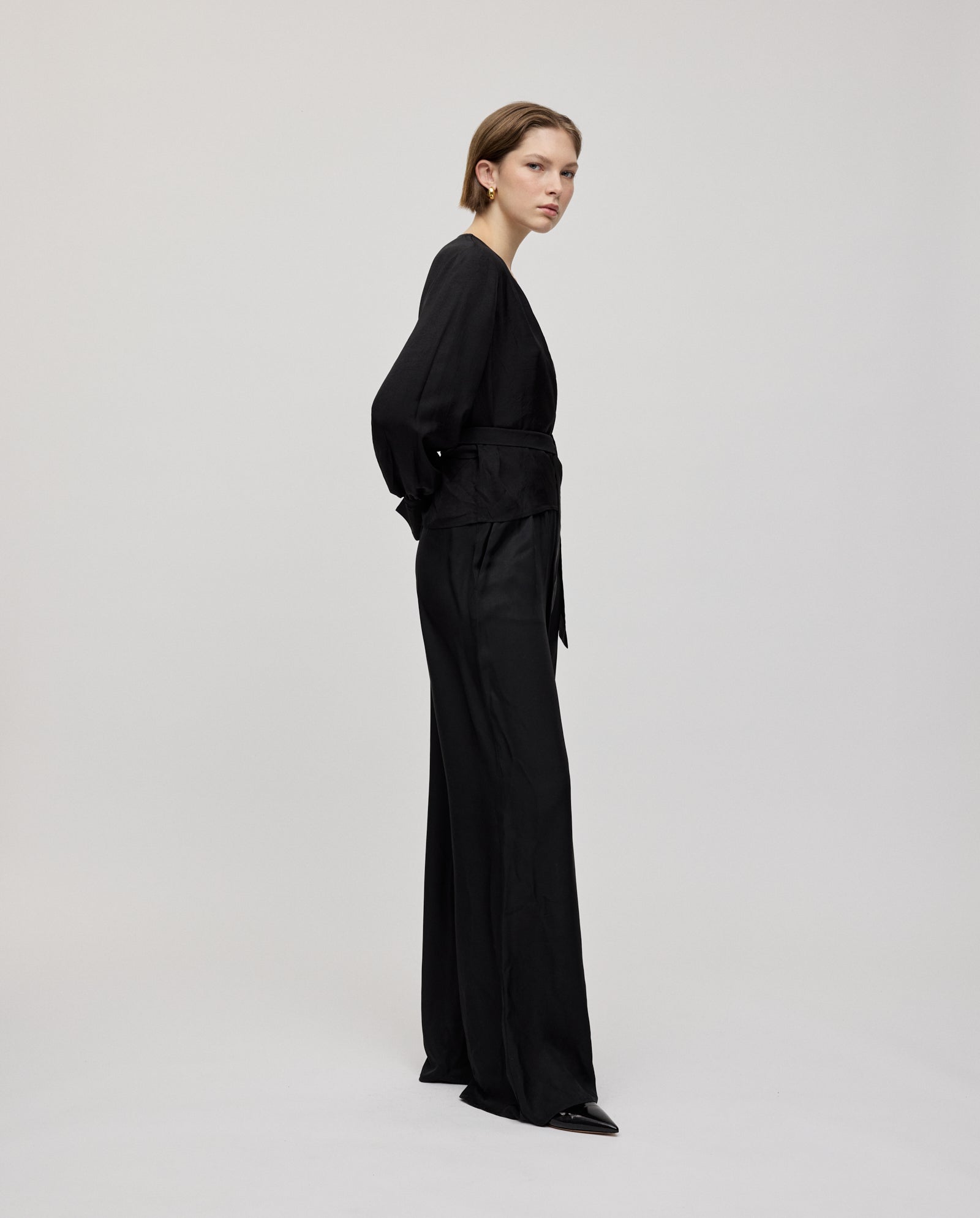 A woman stands sideways against a plain light background, wearing the IVY OAK ELOISE Top—a black organza wrap blouse with long cuffs—paired with wide-leg trousers.