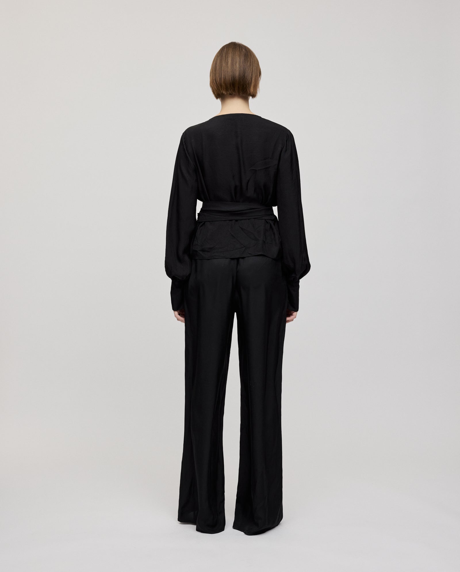 A person with short hair is seen from behind, wearing the IVY OAK ELOISE Top—a black wrap blouse with long cuffs—posing against a plain light background.
