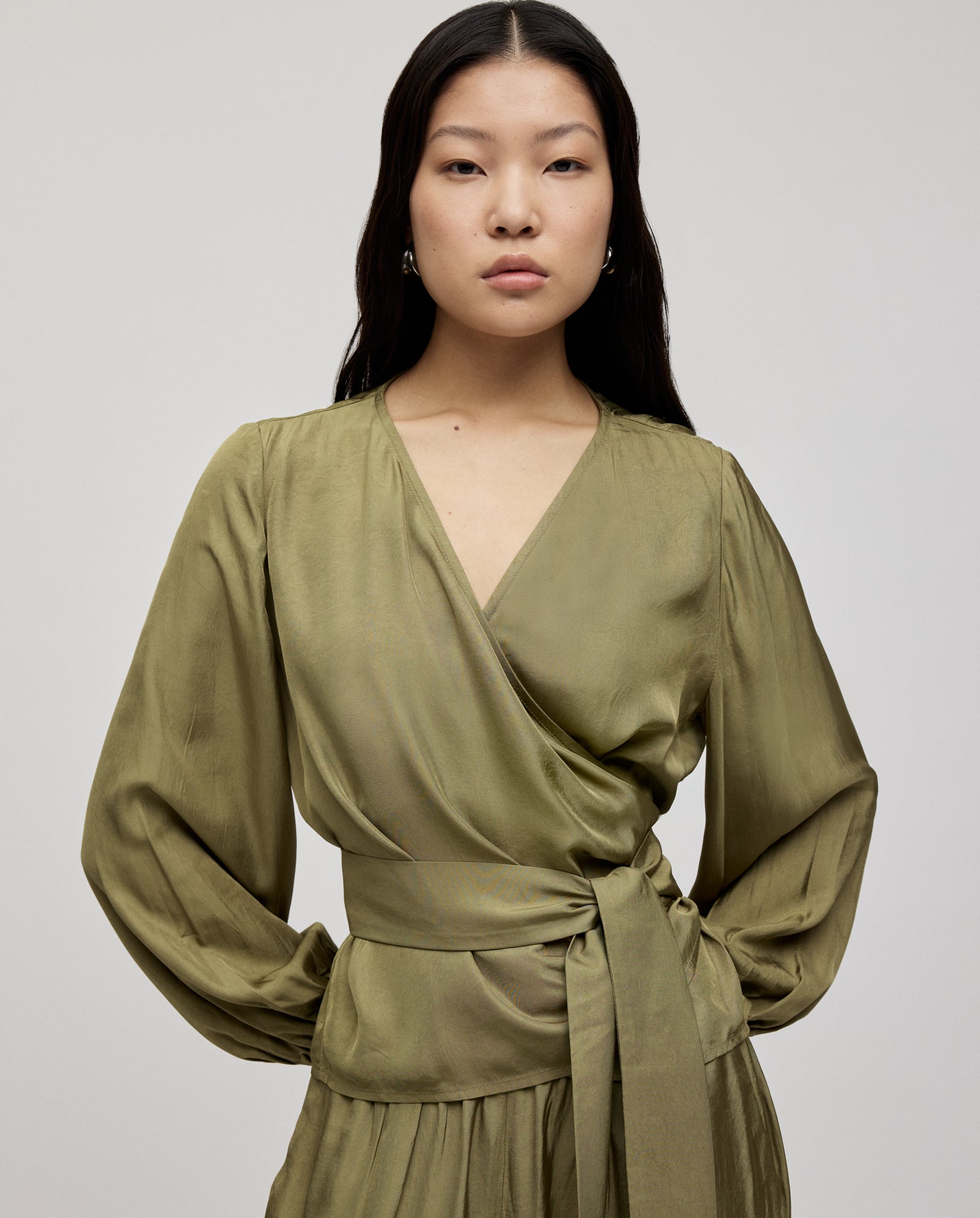 A woman stands against a plain light background wearing the IVY OAK ELOISE Top in silky olive green, styled with a long belt.