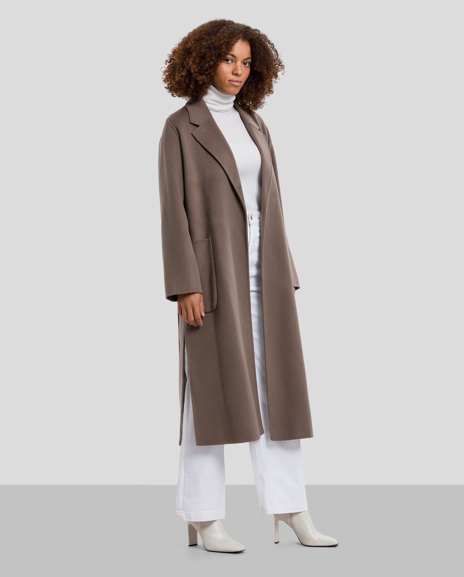 A woman wears the IVY OAK CELIA Doubleface Coat, a taupe midi wool coat made with Responsible Wool Standard materials, paired with a white turtleneck, white pants, and white heeled boots against a gray background.