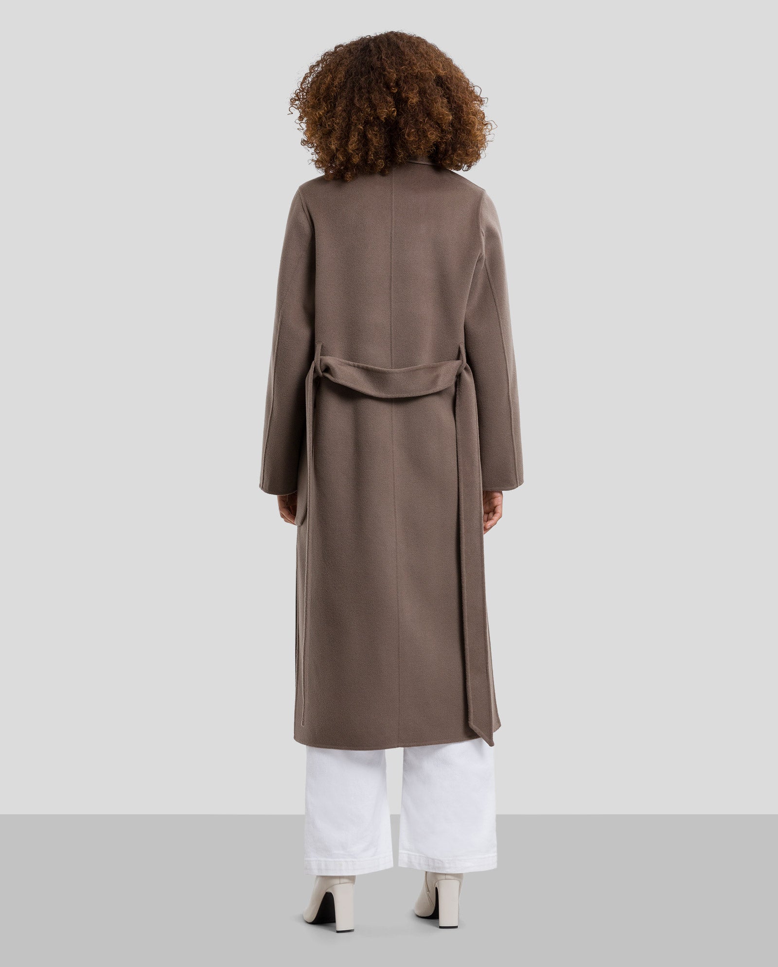A person wearing the IVY OAK CELIA Doubleface Coat in taupe—a midi-length, belted wool coat meeting the Responsible Wool Standard—paired with white pants and heeled boots, seen from the back.