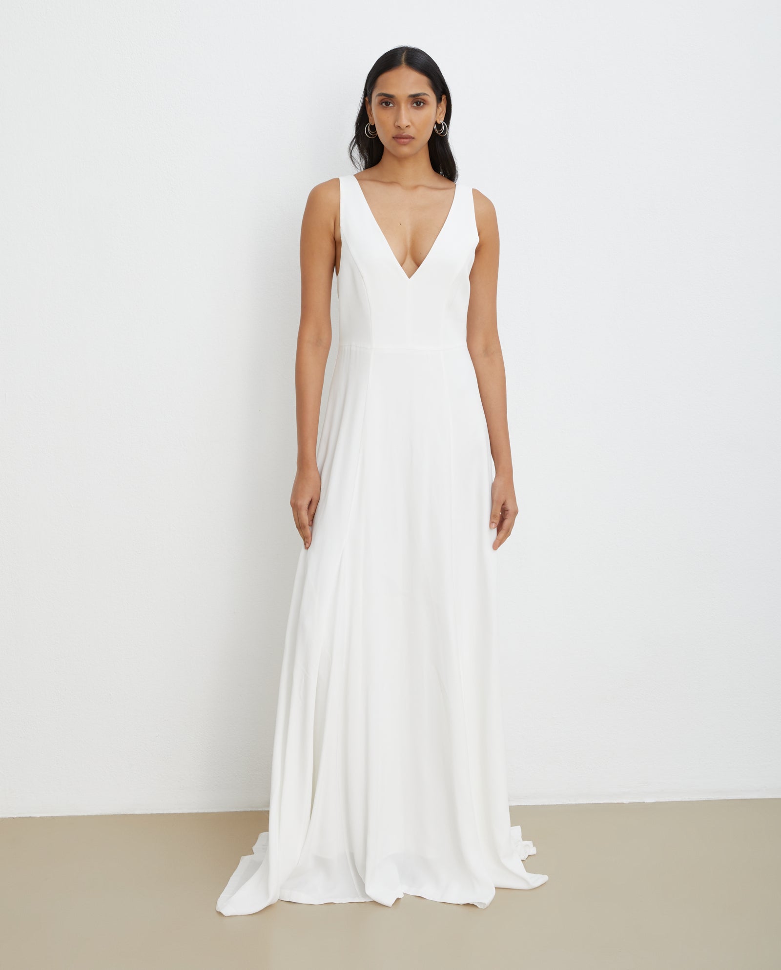A woman in the IVY OAK STEVIA Bridaldress, a sleeveless, floor-length viscose gown with a deep V-neckline, stands against a plain wall.