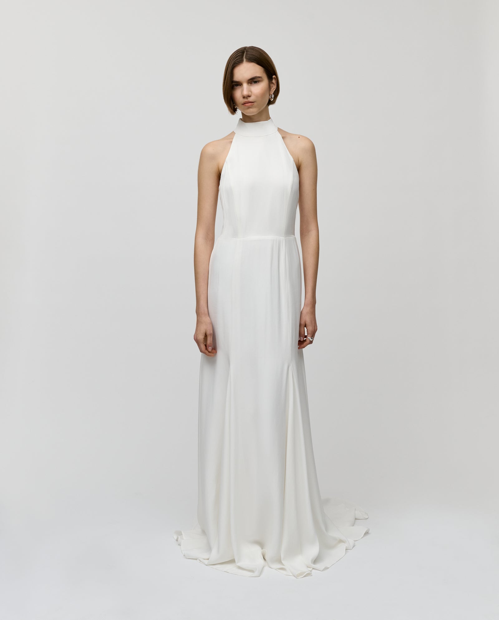 A woman wears the IVY OAK MEREDITH Dress—a sleeveless, high-neck, maxi-length halter bridal gown in crepe—standing against a plain white background.