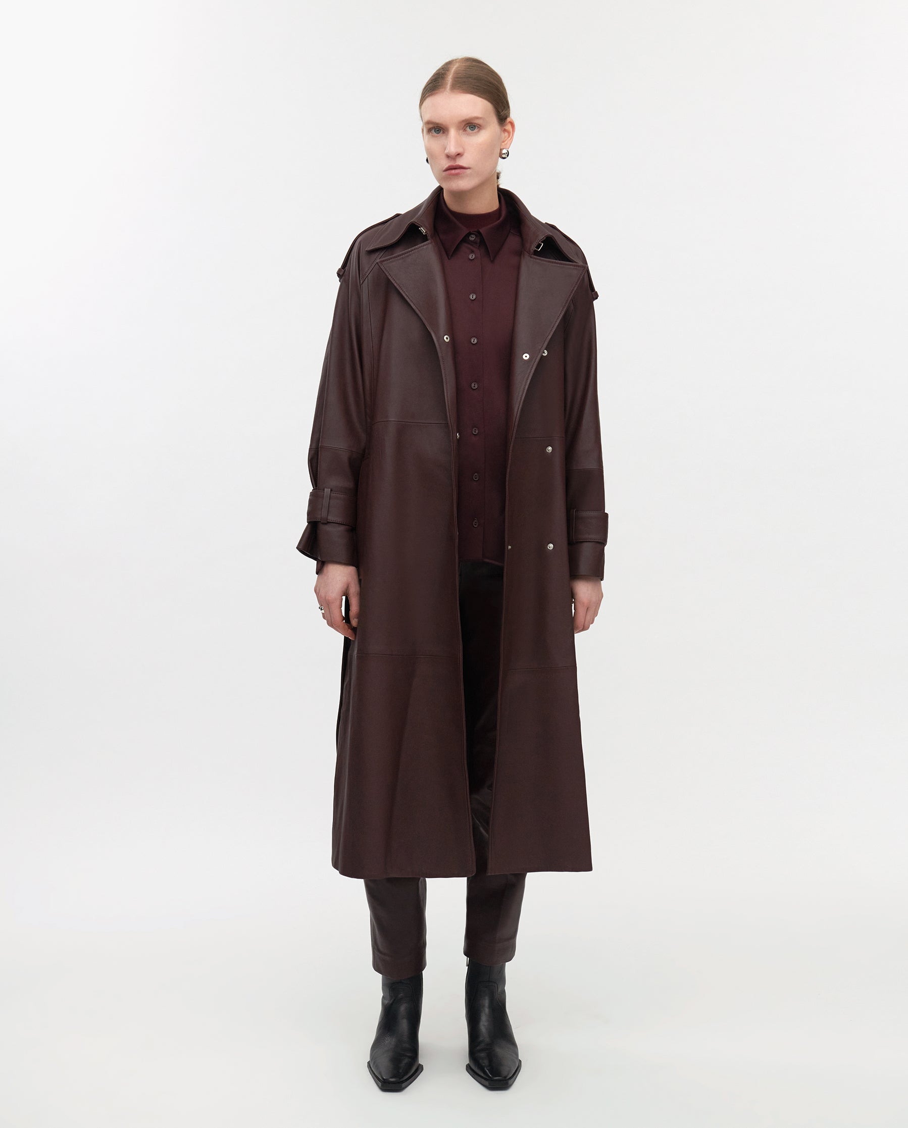 A person wears the IVY OAK LILITH Trench, a long, dark brown belted coat, over matching pants and shirt, standing against a white background.
