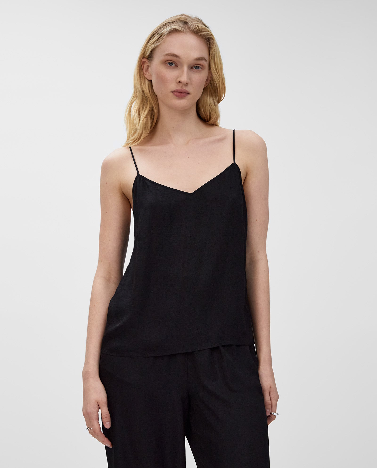 A woman with long blonde hair wears a black IVY OAK TILDA ANN top featuring a V-neckline and black pants, standing against a plain background.