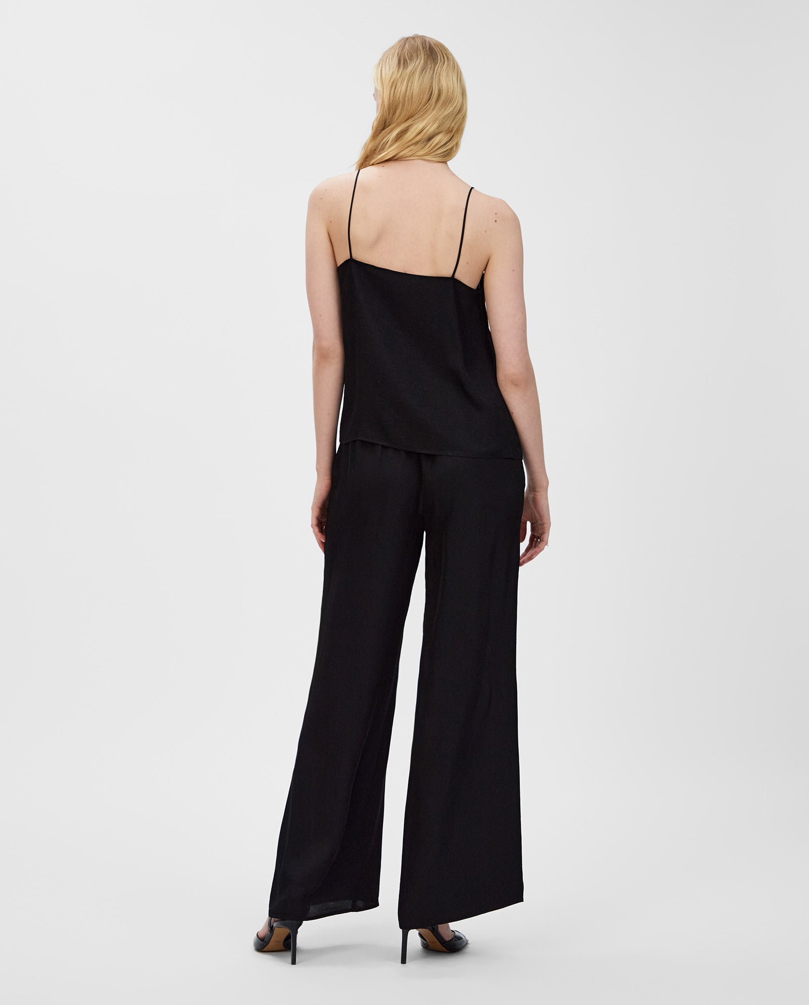 A woman with blonde hair wears the IVY OAK TILDA ANN Top, a black shiny V-neck camisole, paired with wide-leg pants, standing with her back to the camera.