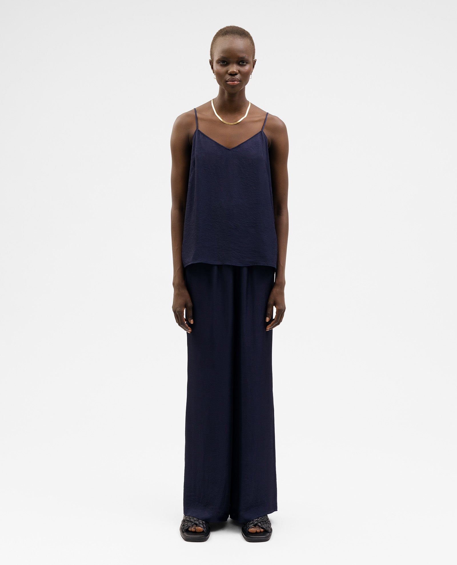 A person wears a navy IVY OAK TILDA ANN Top with a V-neckline, wide-leg pants, sandals, and a necklace, standing against a plain background.