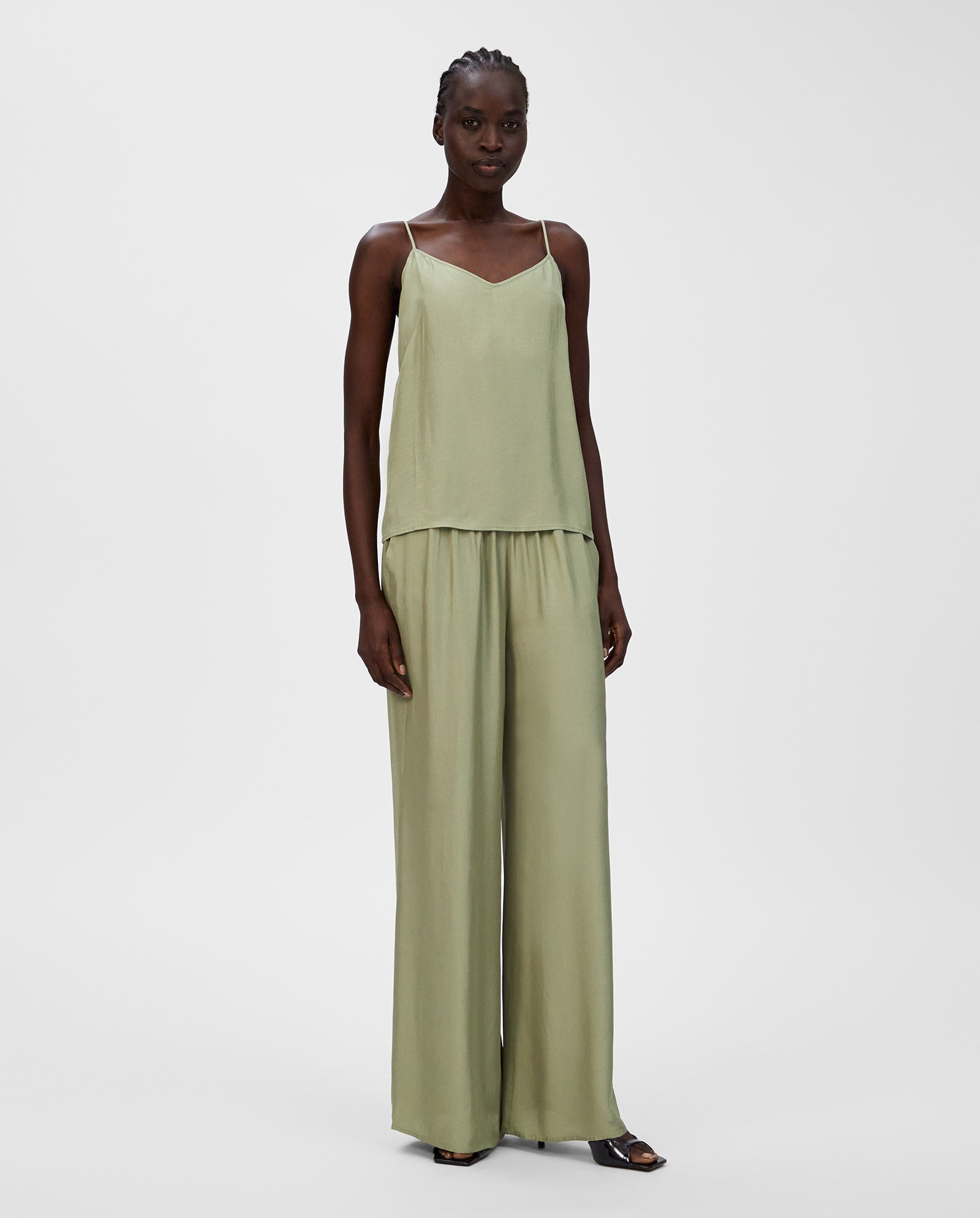A woman wears a light green, sleeveless, loose-fit TILDA ANN Top by IVY OAK with a V-neckline and matching wide-leg pants made from soft Naia acetate.