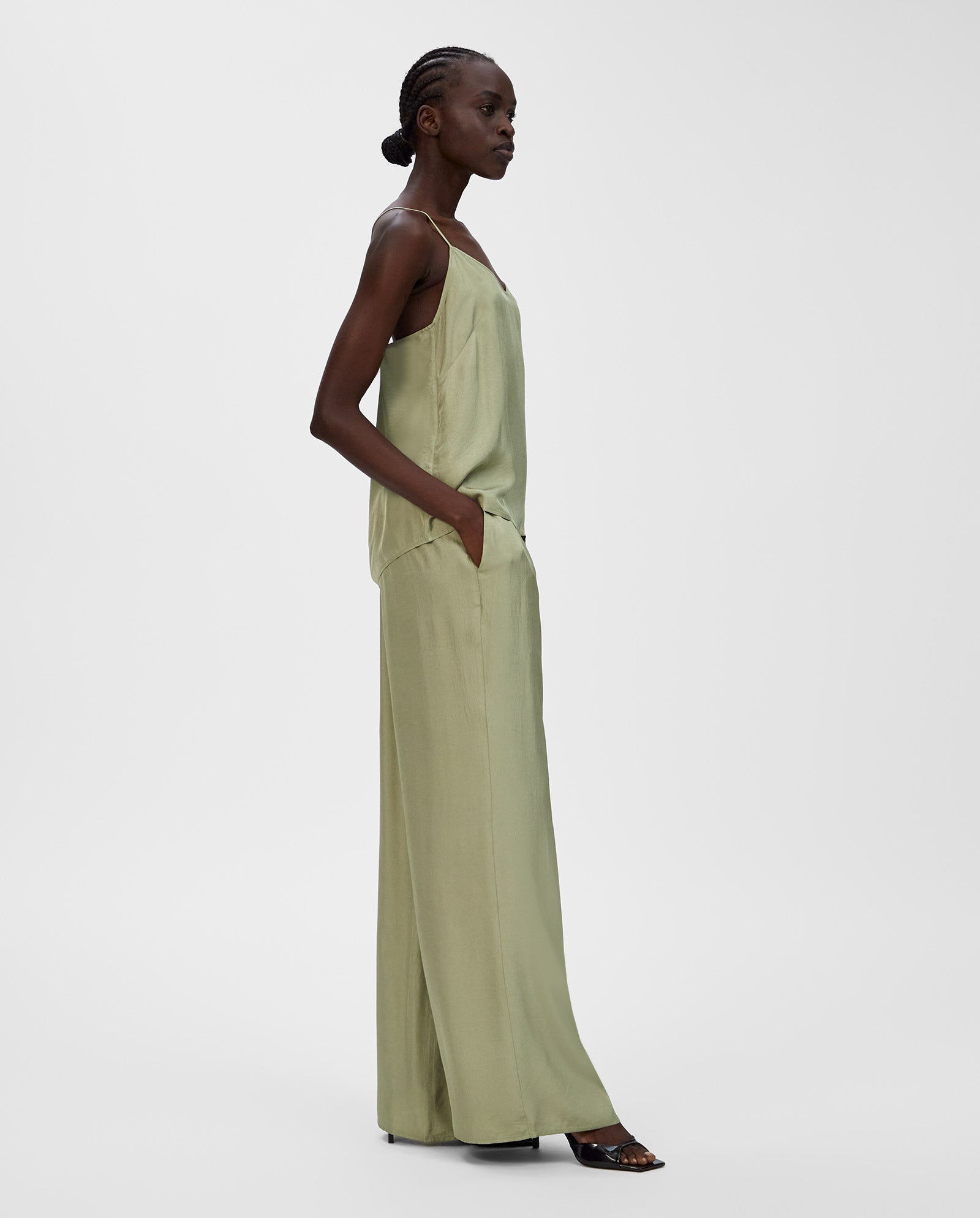 Woman in profile wears the IVY OAK TILDA ANN Top, a light green sleeveless V-neck made from Naia acetate, paired with wide-leg pants and hands in pockets.