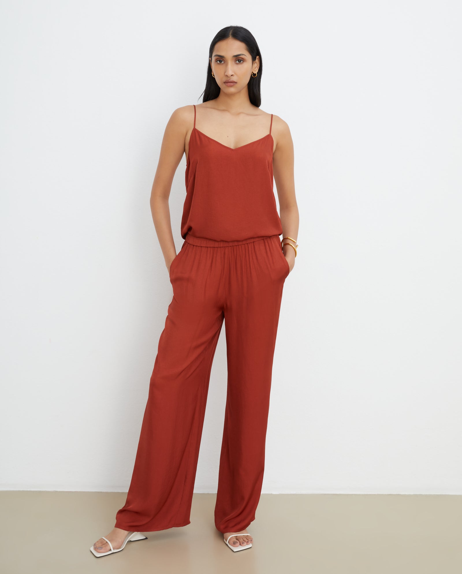 A woman wears the IVY OAK TILDA ANN rust-colored spaghetti strap top with a V-neckline, paired with white sandals, standing against a white wall.