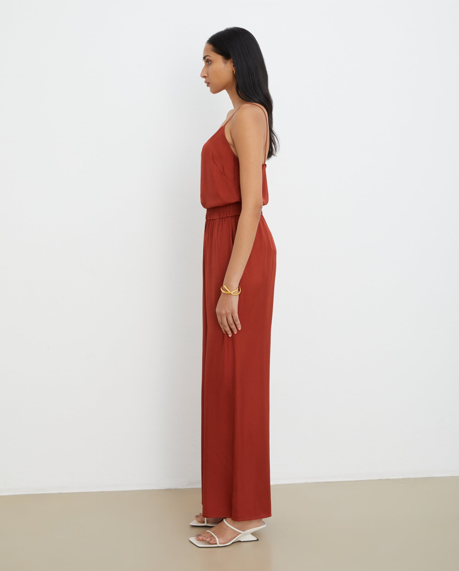 Woman wearing the IVY OAK TILDA ANN Top, a sleeveless rust-colored V-neck, paired with sandals and standing in profile against a plain white wall.