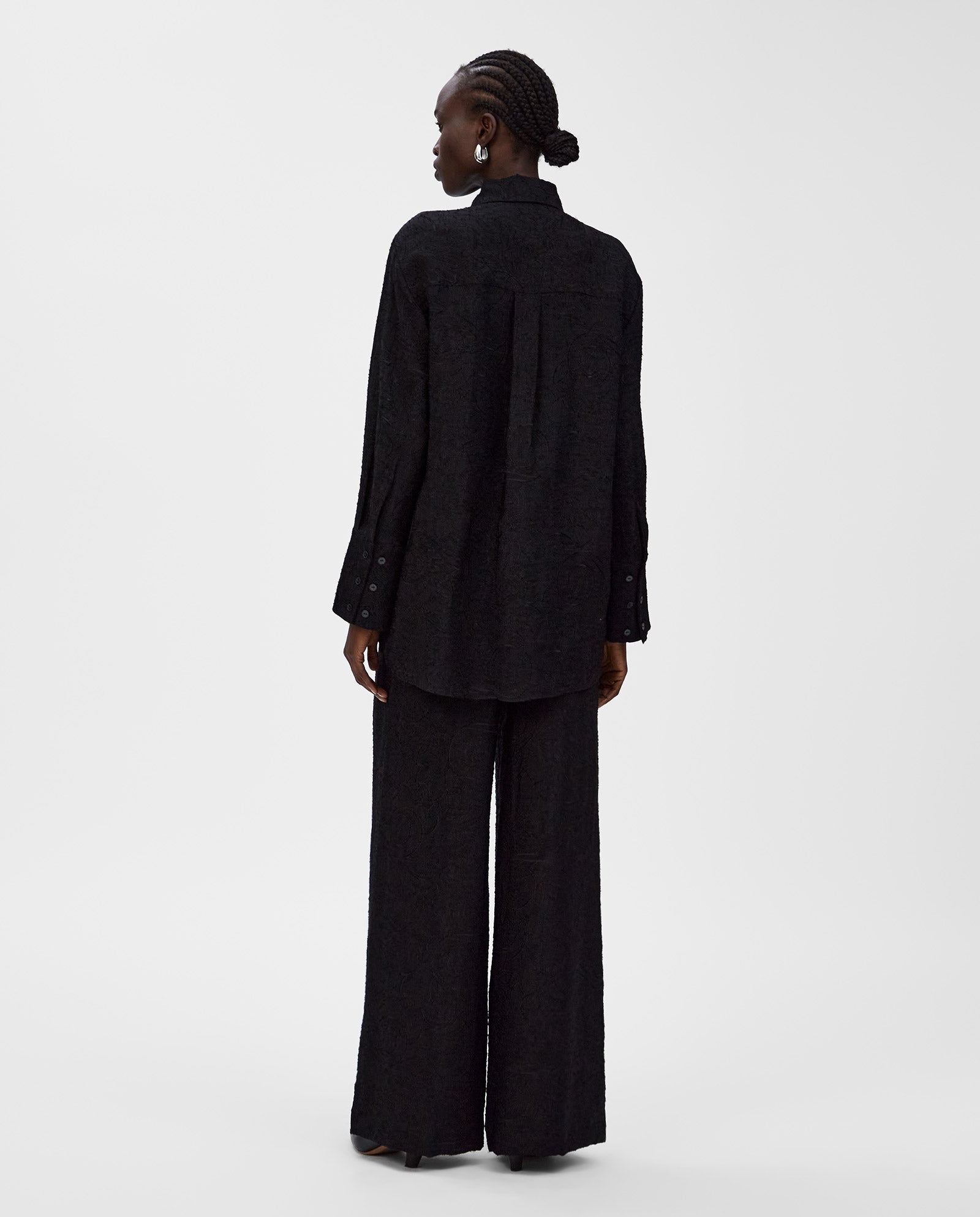 A person is seen from the back wearing the IVY OAK ELVIE ANN Blouse, a loose black fil coupé design with a wide collar, paired with wide-leg pants.