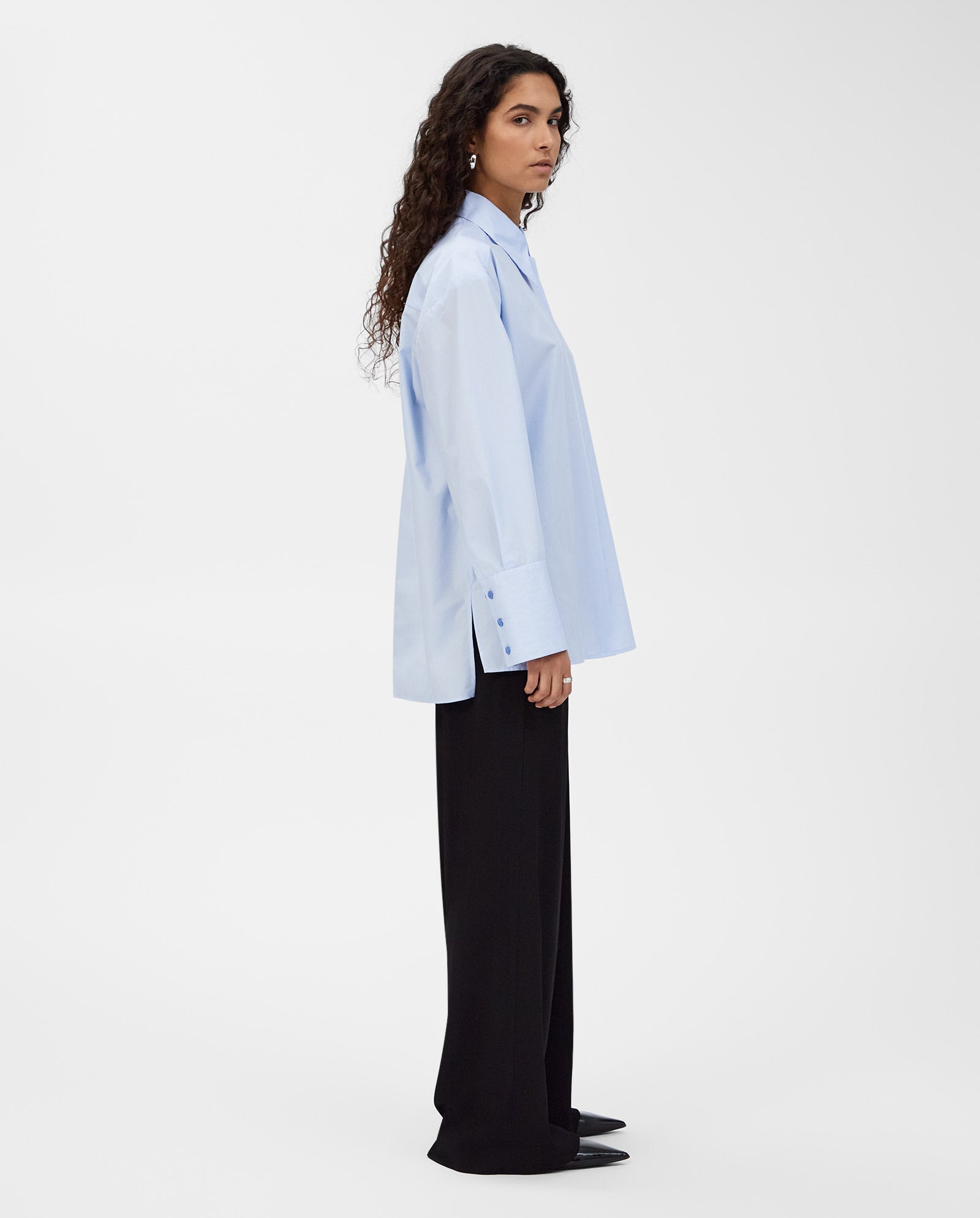 A woman stands sideways against a plain white background, wearing the IVY OAK ELVIE Blouse—a loose, light blue shirt with a wide collar—paired with black pants for a modern classic look.