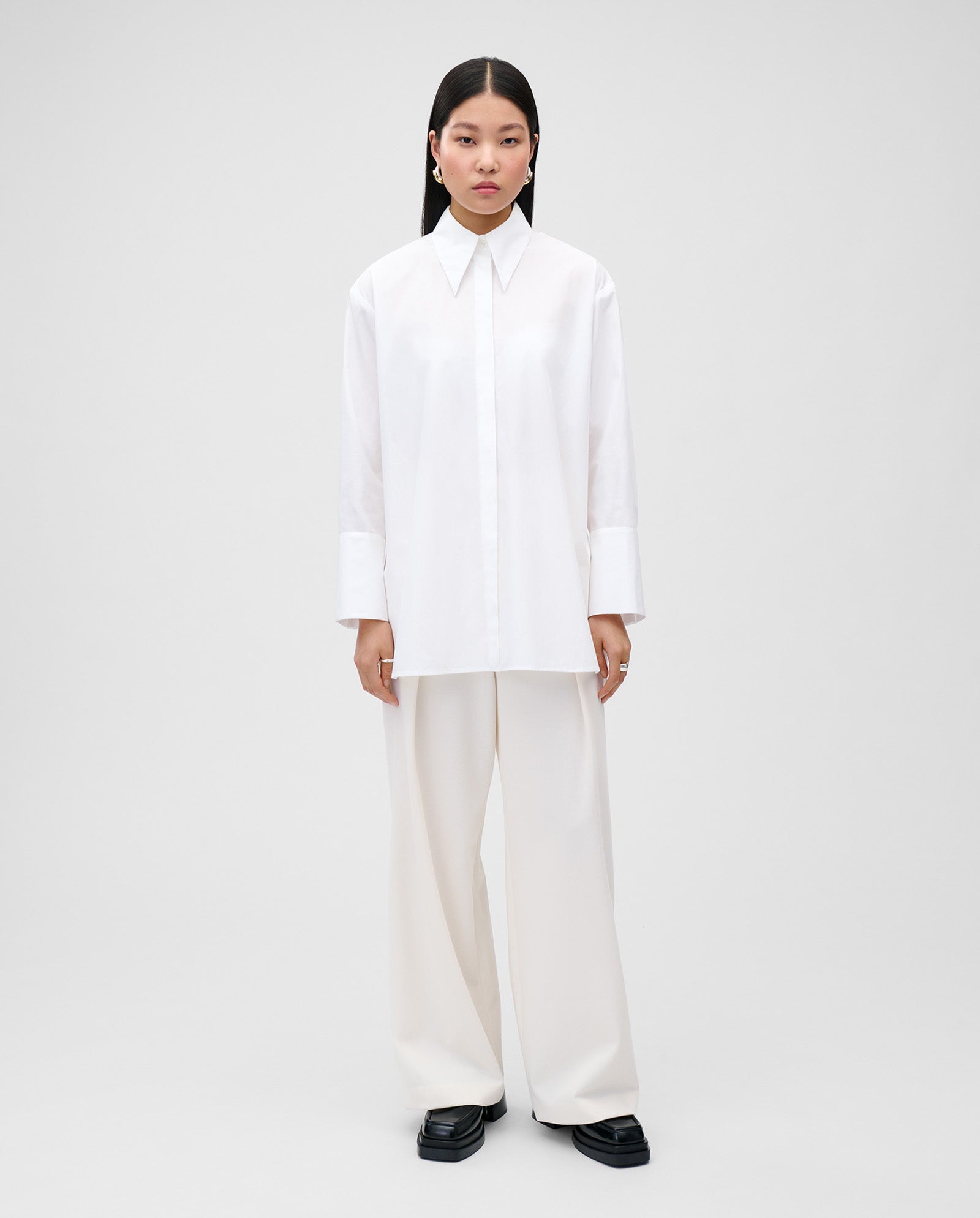 A woman models the IVY OAK ELVIE Blouse, featuring adjustable cuffs and a wide collar, styled with loose white pants and black shoes against a plain background.