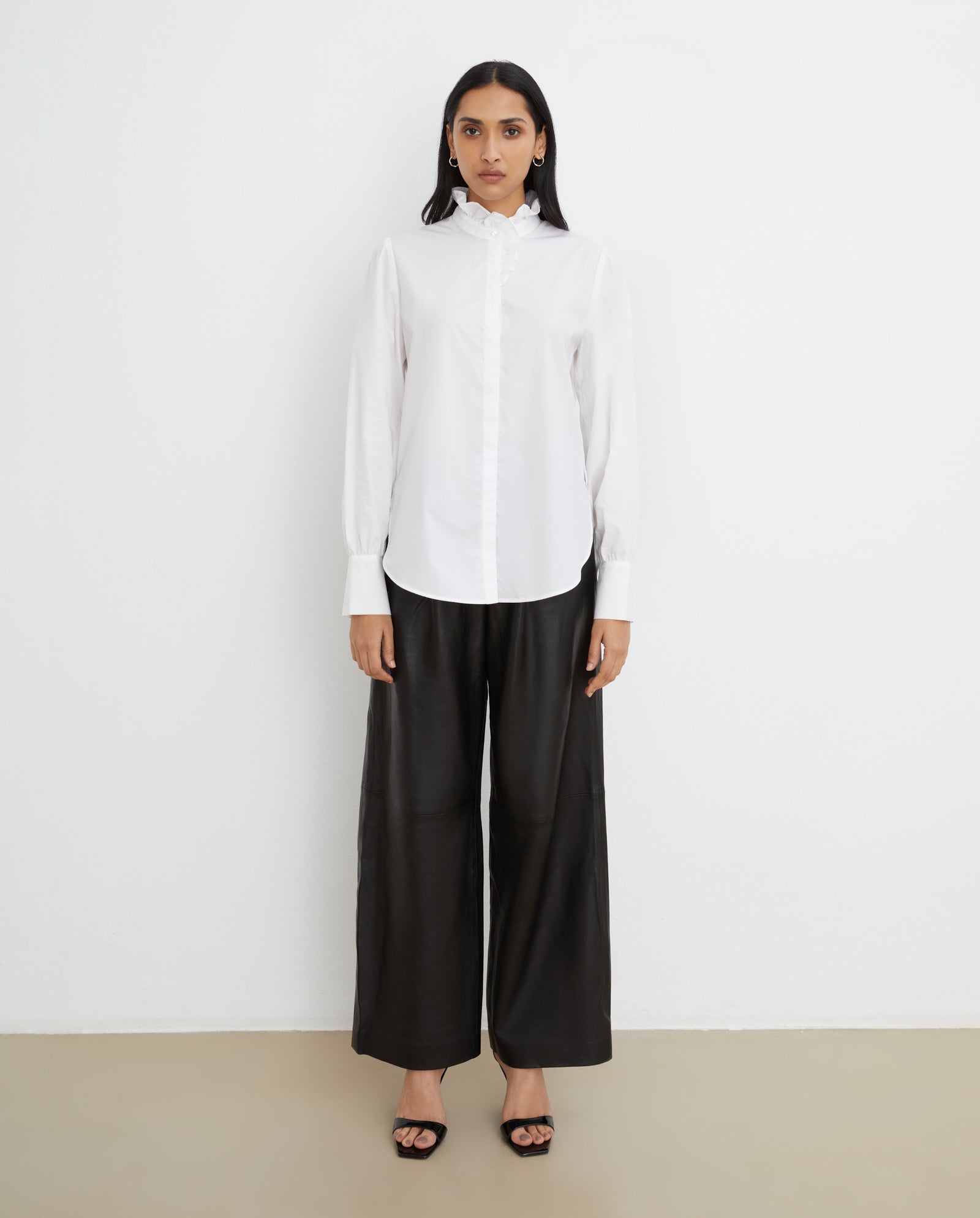 A woman wears the IVY OAK ESTELLE Top, featuring a white ruffled stand-up collar and hidden button placket, paired with black wide-leg pants, standing against a plain white wall.