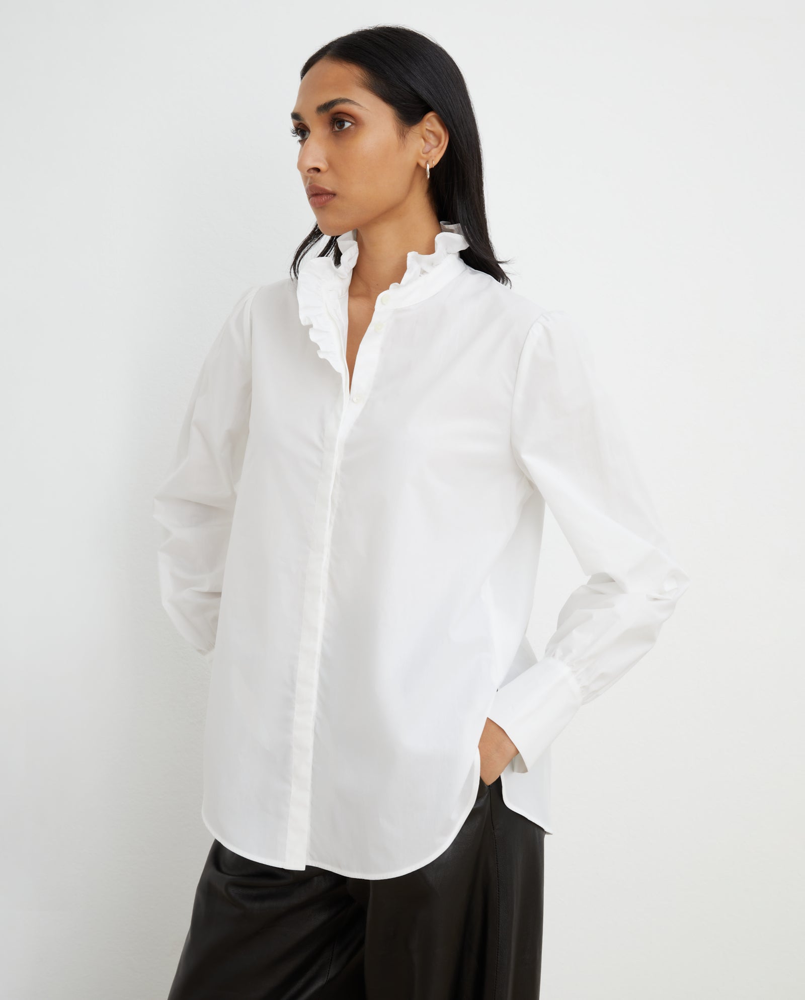 A woman wears the ESTELLE Top by IVY OAK, featuring a ruffled stand-up collar and hidden button placket, styled with black pants and posed against a plain white background.