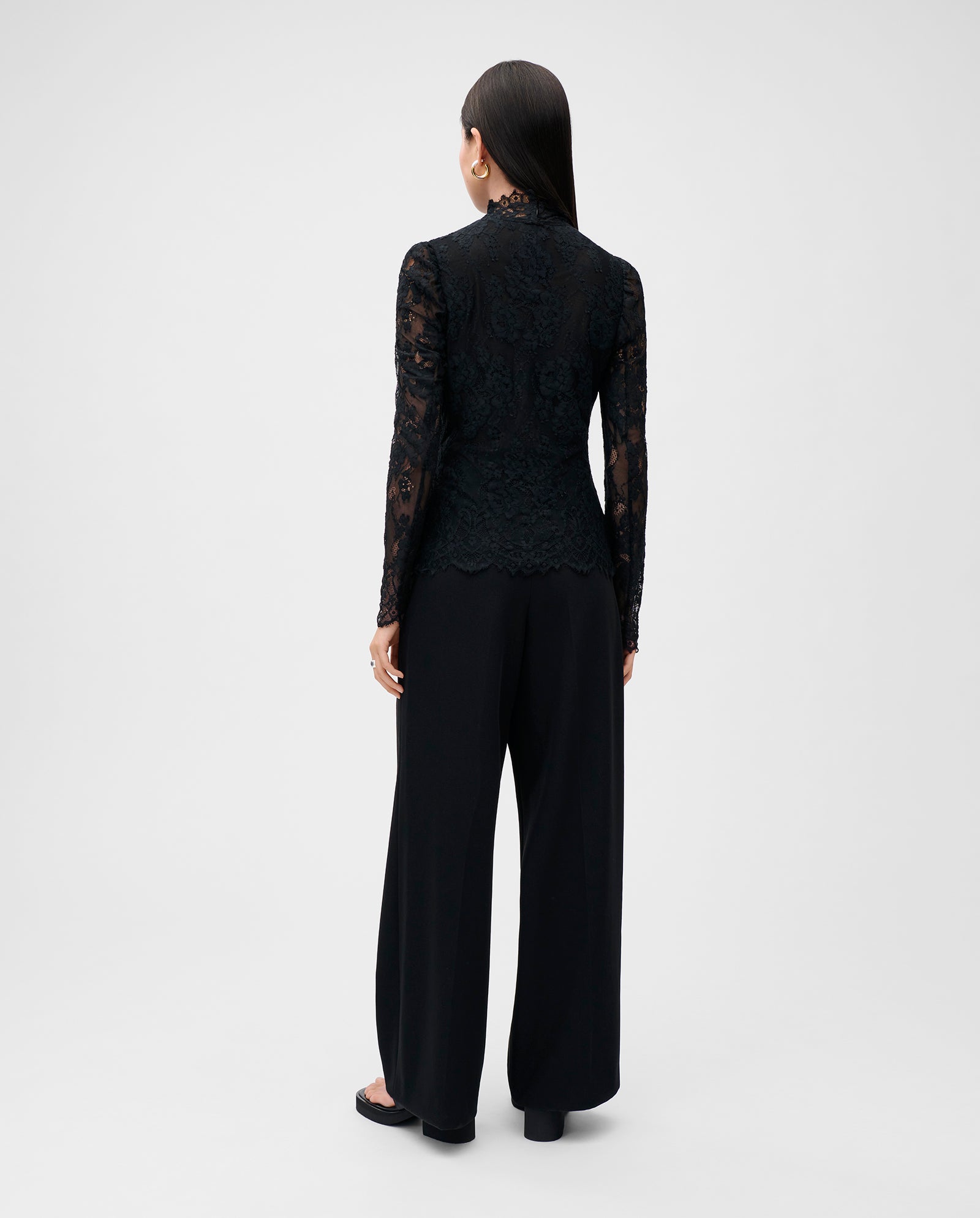 A woman is seen from behind wearing the IVY OAK BRIHANNA Top, a black lace long-sleeve blouse, paired with wide-leg black pants for an elegant look.
