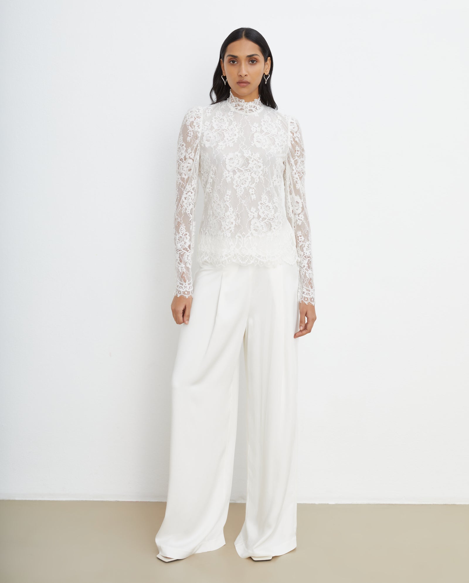 A woman models the IVY OAK BRIHANNA Top—a white, sustainable lace long-sleeve blouse—paired with wide-leg white trousers, standing against a plain background for an elegant look.