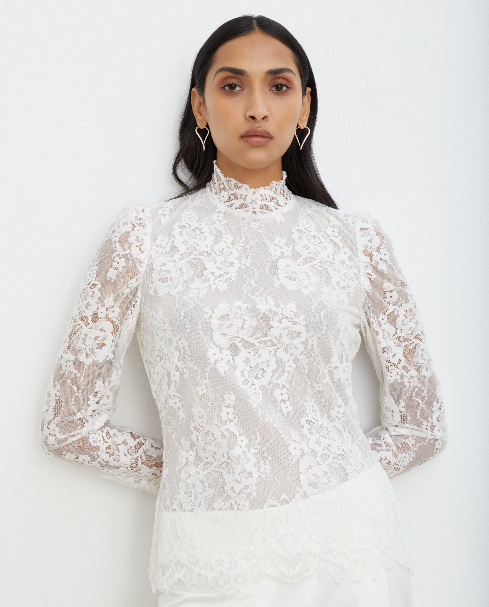 Model wears the IVY OAK BRIHANNA Top—a white lace long-sleeve piece with sheer floral details and mesh lining—standing against a white background for an elegant look.
