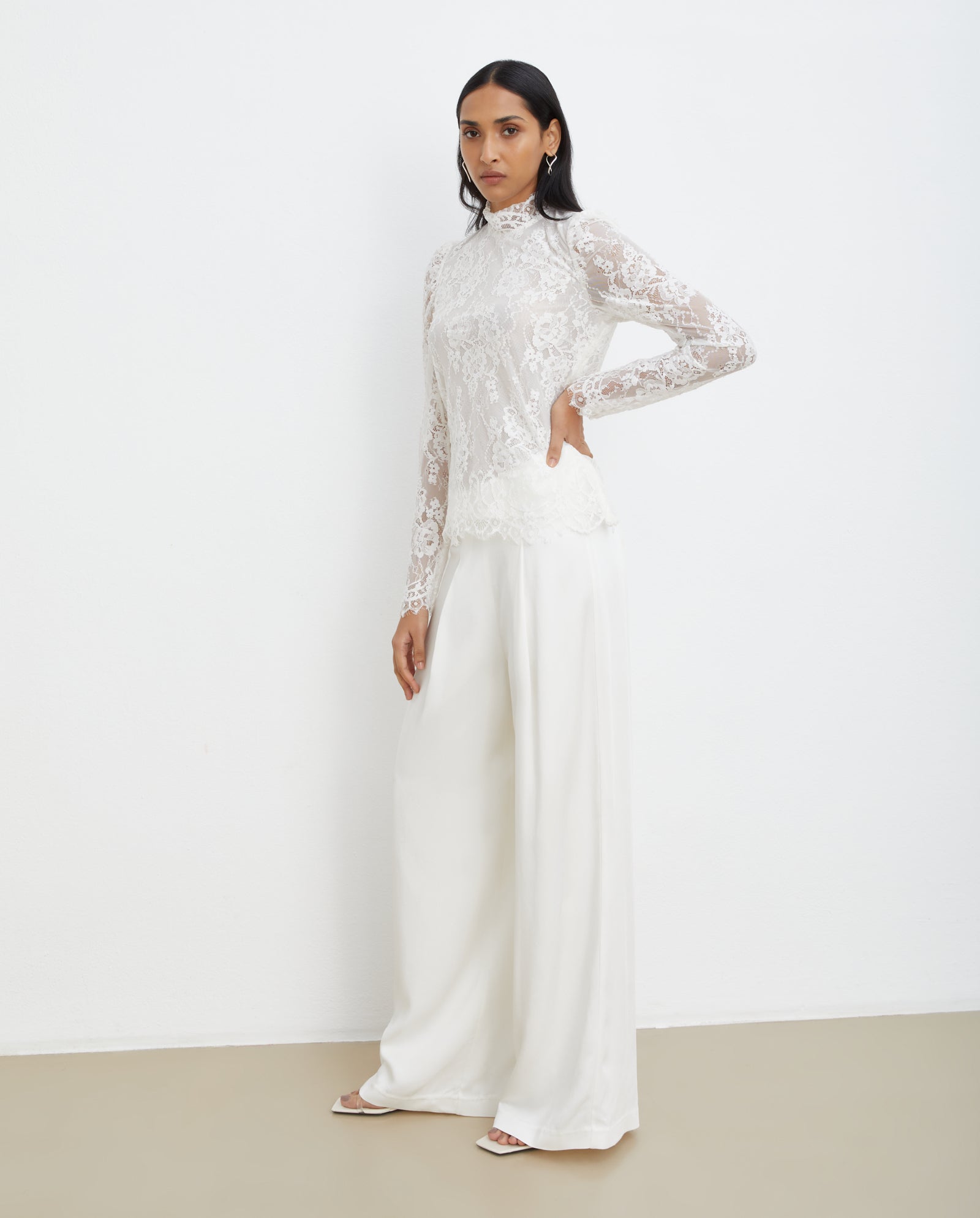A woman wears the BRIHANNA Top by IVY OAK—a white lace, long-sleeve design with mesh lining—paired with wide-leg pants, posing against a plain white wall for an elegant look.