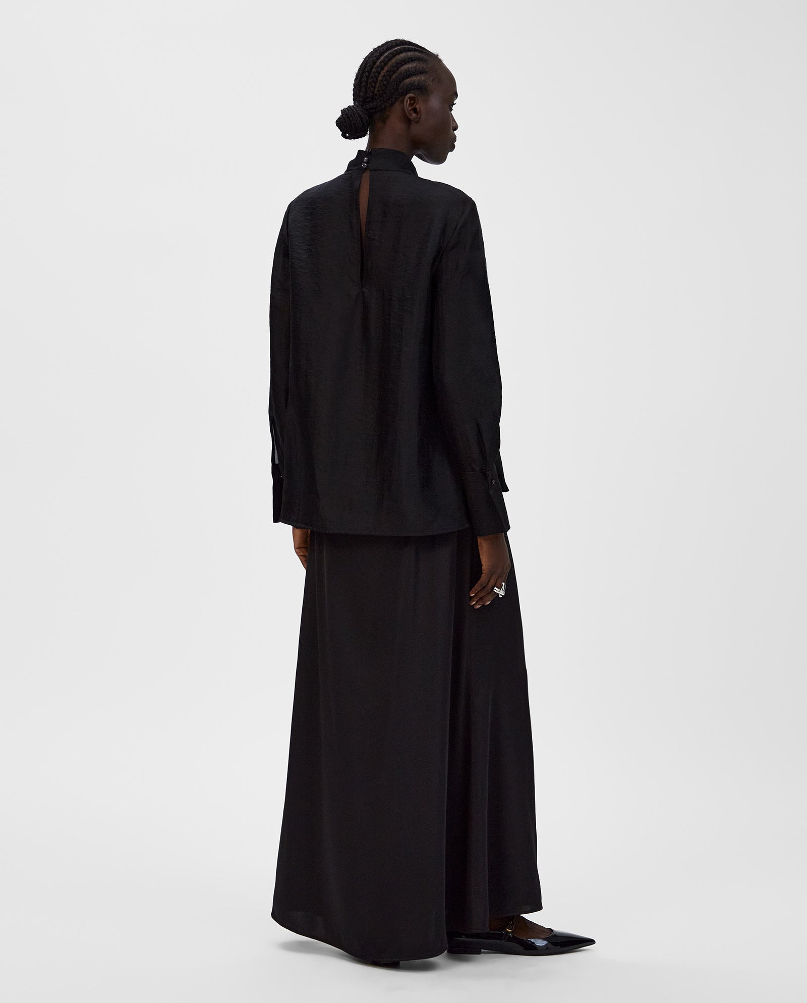 A person with braided hair wears a loose black IVY OAK EBONEY Blouse and a long black skirt, seen from behind on a plain background.