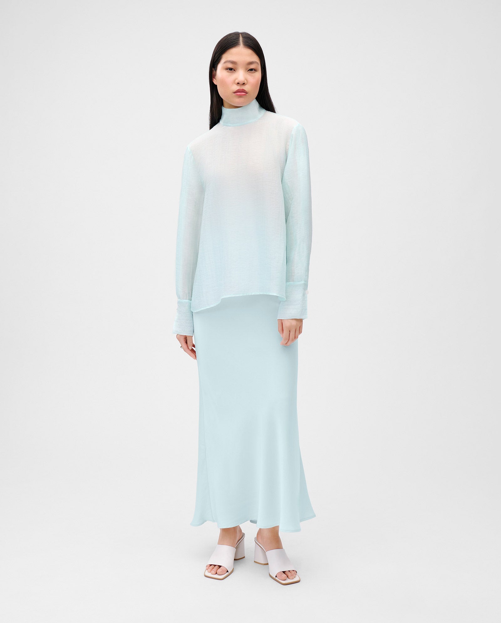 A woman wears the IVY OAK EBONEY Blouse, an elegant light blue organza stand-up collar turtleneck top, paired with a matching long skirt, against a plain background.