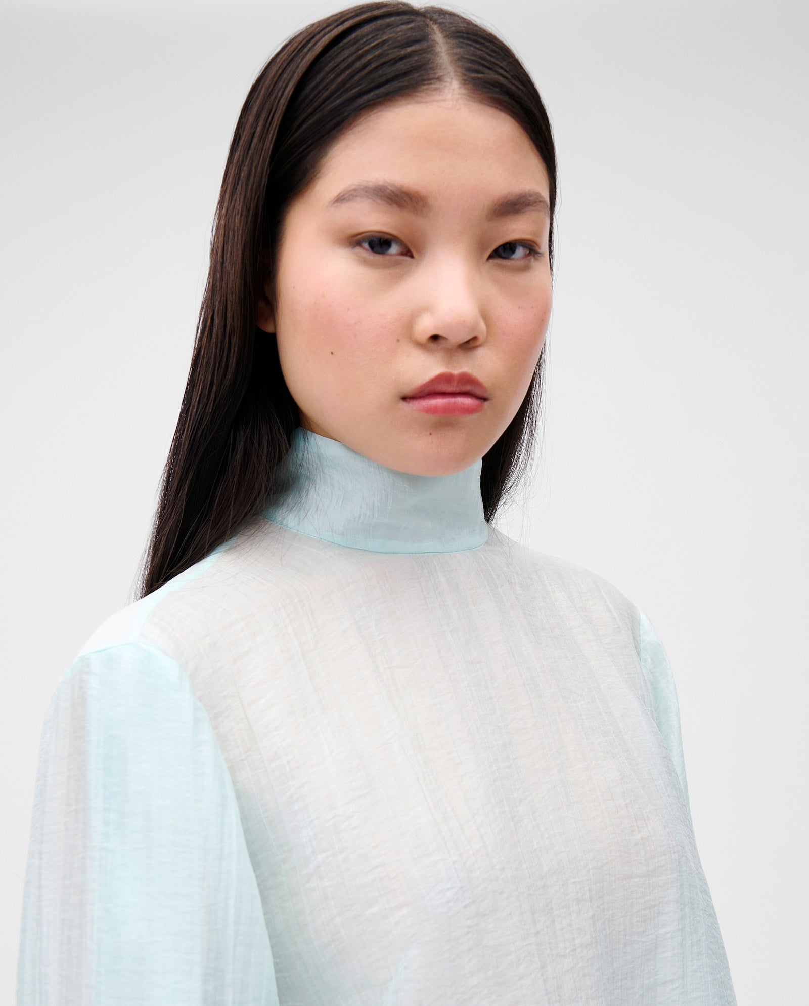 A woman with straight dark hair wears the IVY OAK EBONEY Blouse, an elegant light blue organza top with a high stand-up collar, and looks at the camera.