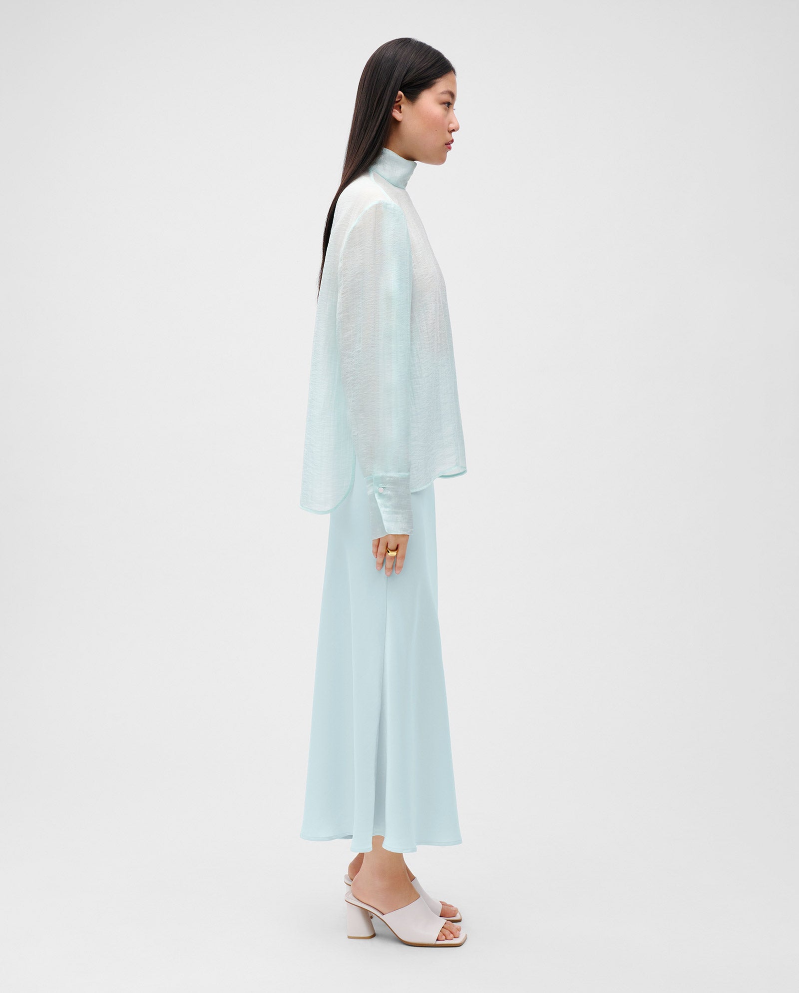 Woman in profile wears the IVY OAK EBONEY Blouse, an elegant light blue sheer organza top with a stand-up collar and skirt, set against a plain white background.