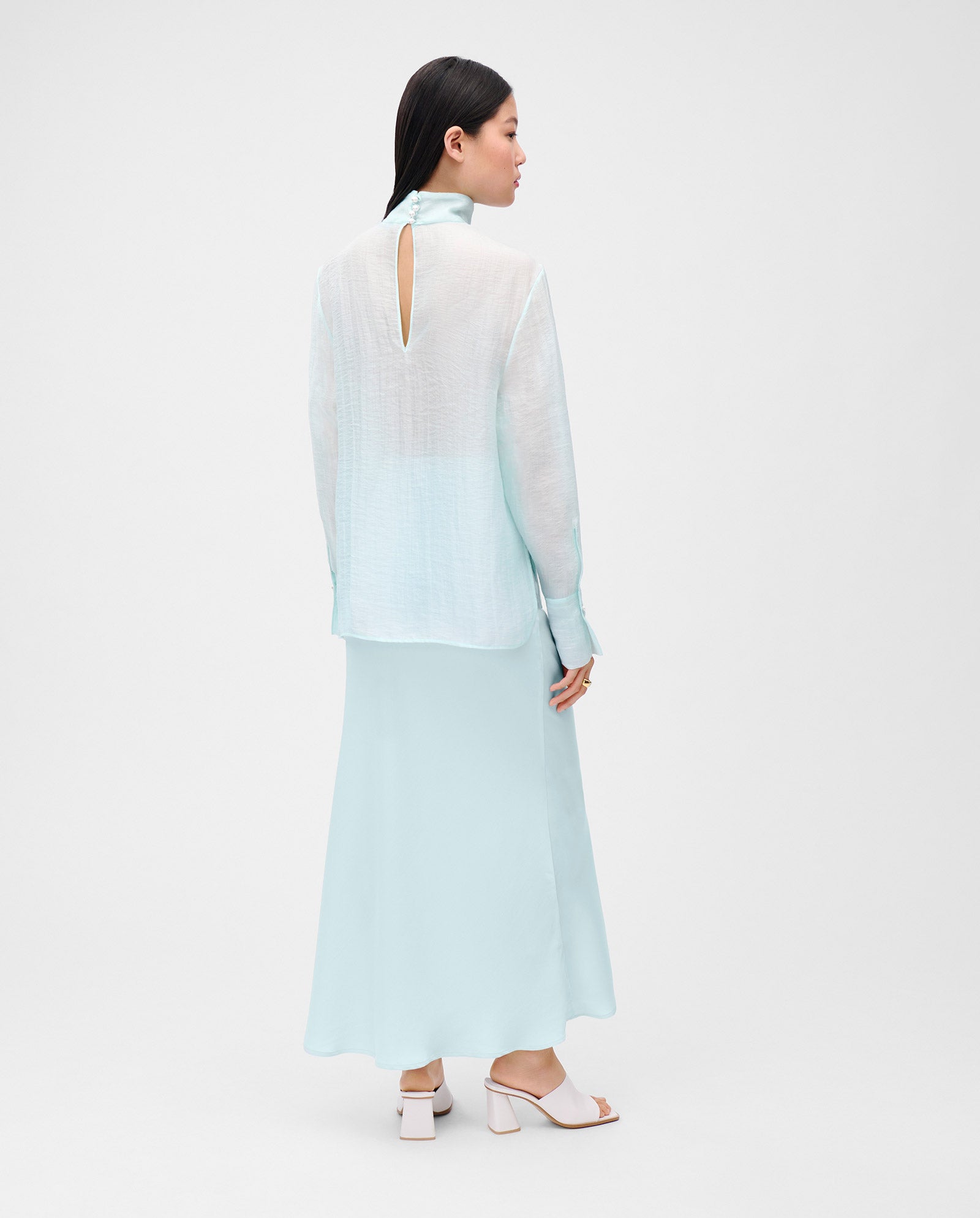 A woman models the IVY OAK EBONEY Blouse, a light blue tunic with a stand-up collar, paired with a matching long skirt, standing sideways against a plain white background.