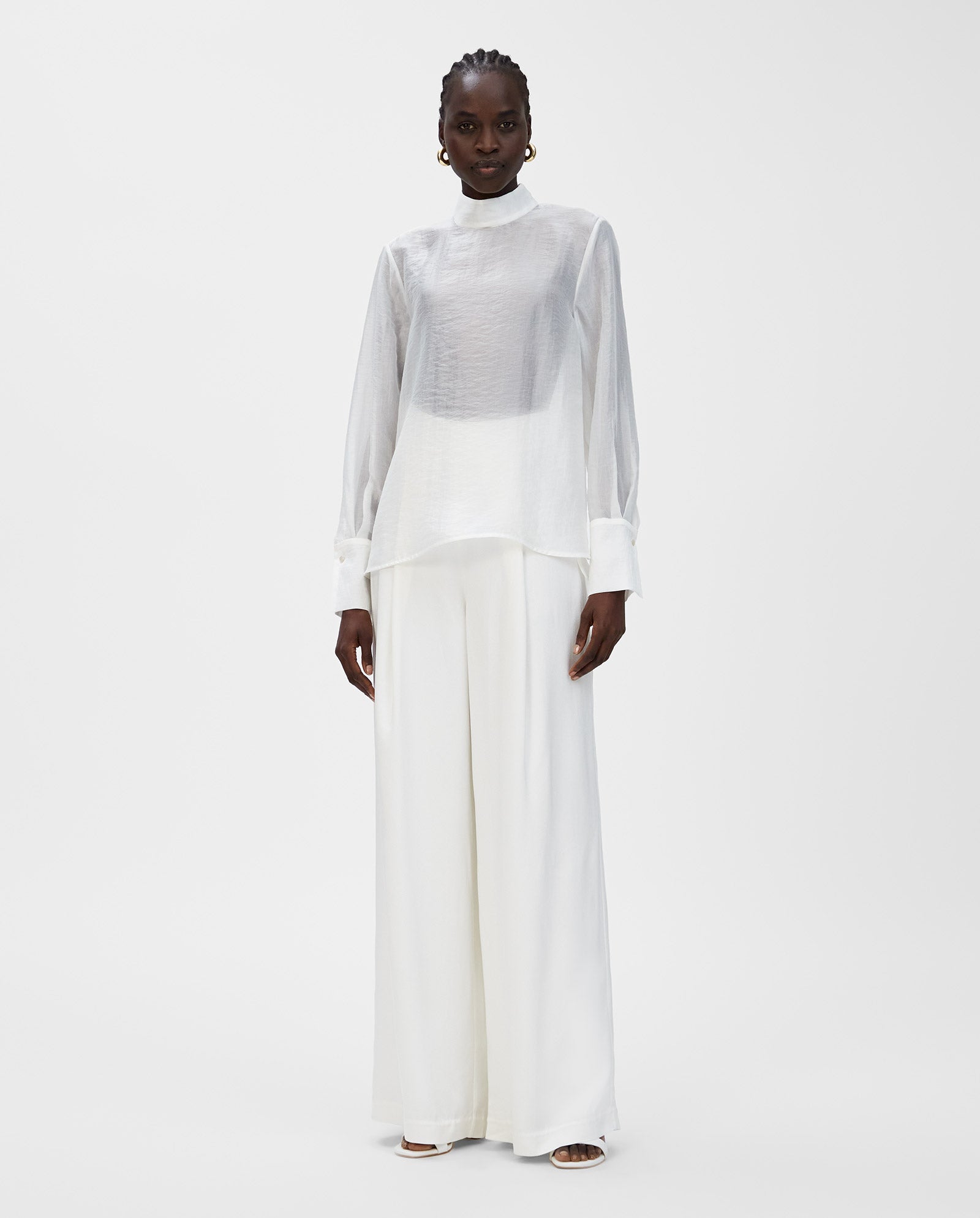 A woman wears the IVY OAK EBONEY Blouse, a sheer organza design with a stand-up collar, paired with wide-leg white trousers against a plain white background, showcasing versatile styling.