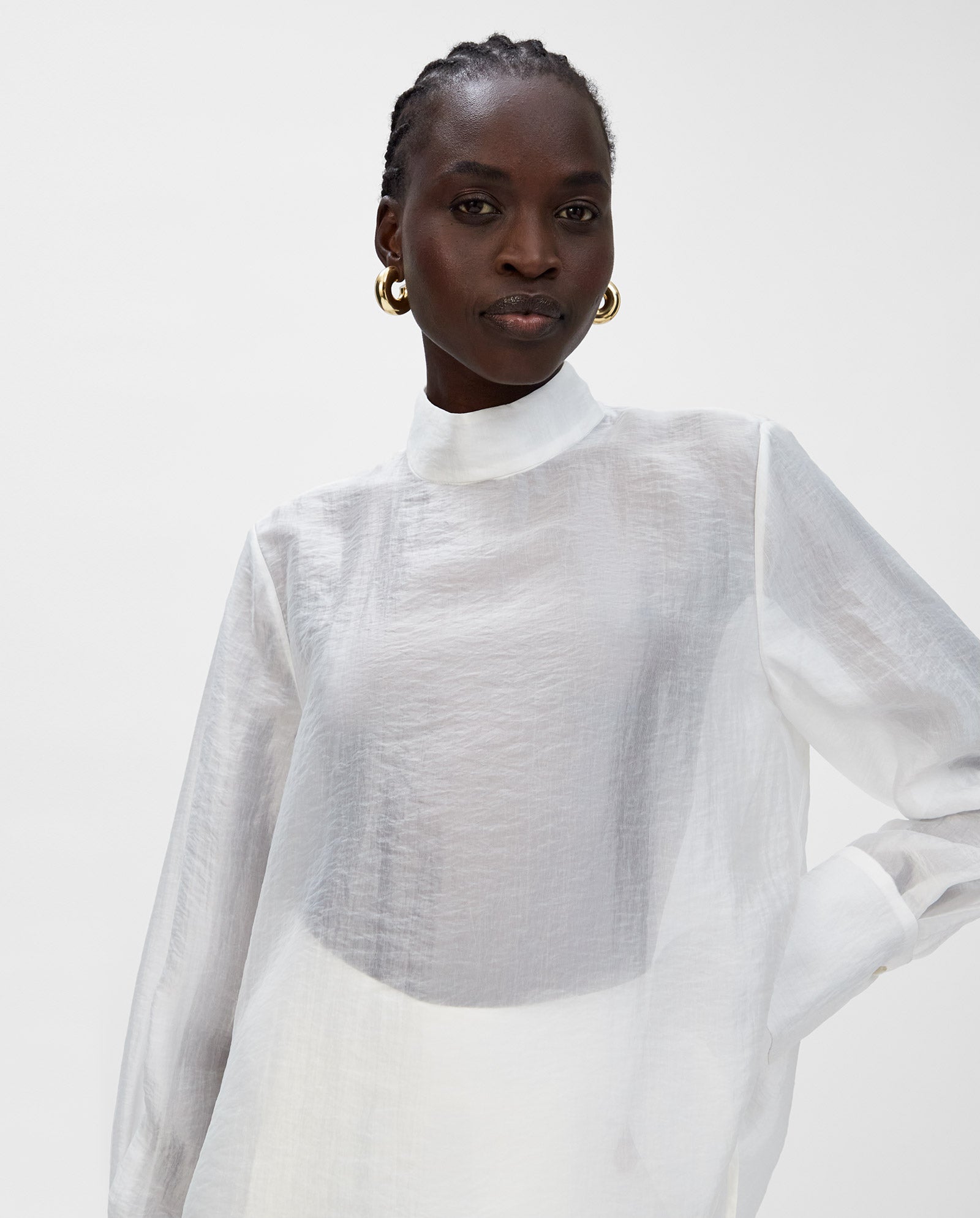 A woman poses against a plain background wearing the IVY OAK EBONEY Blouse, a sheer white organza top with a stand-up collar, styled with gold hoop earrings.