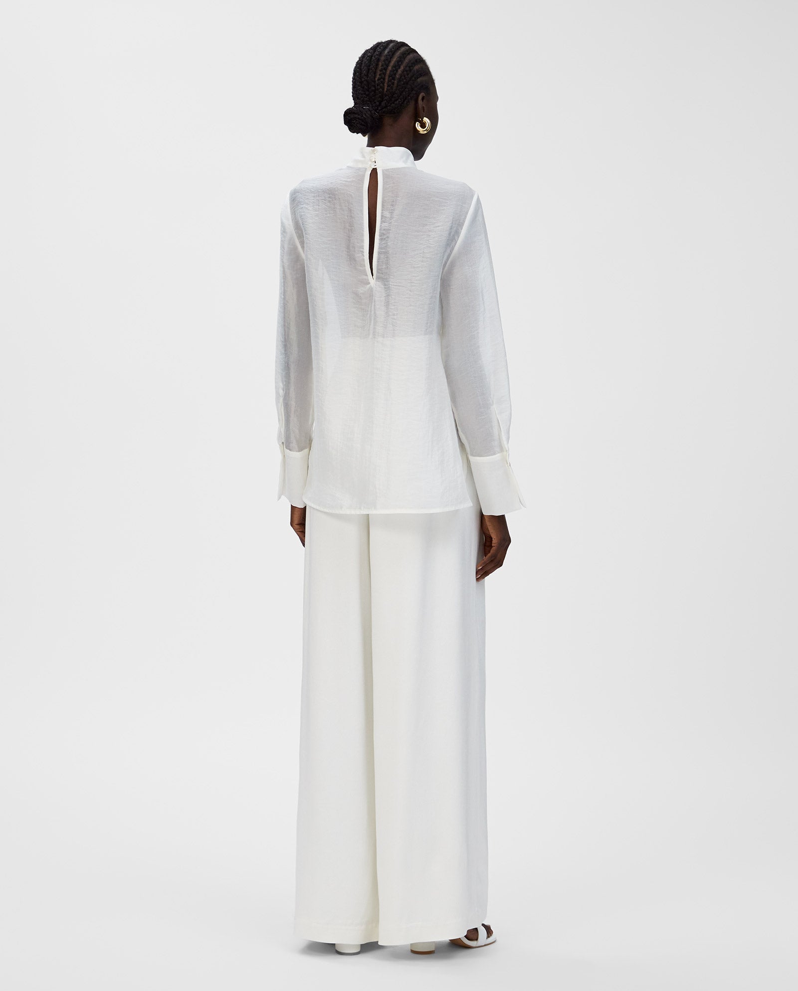 A person faces away against a white background wearing the IVY OAK EBONEY Blouse—white organza with a stand-up collar—paired with wide-leg pants.