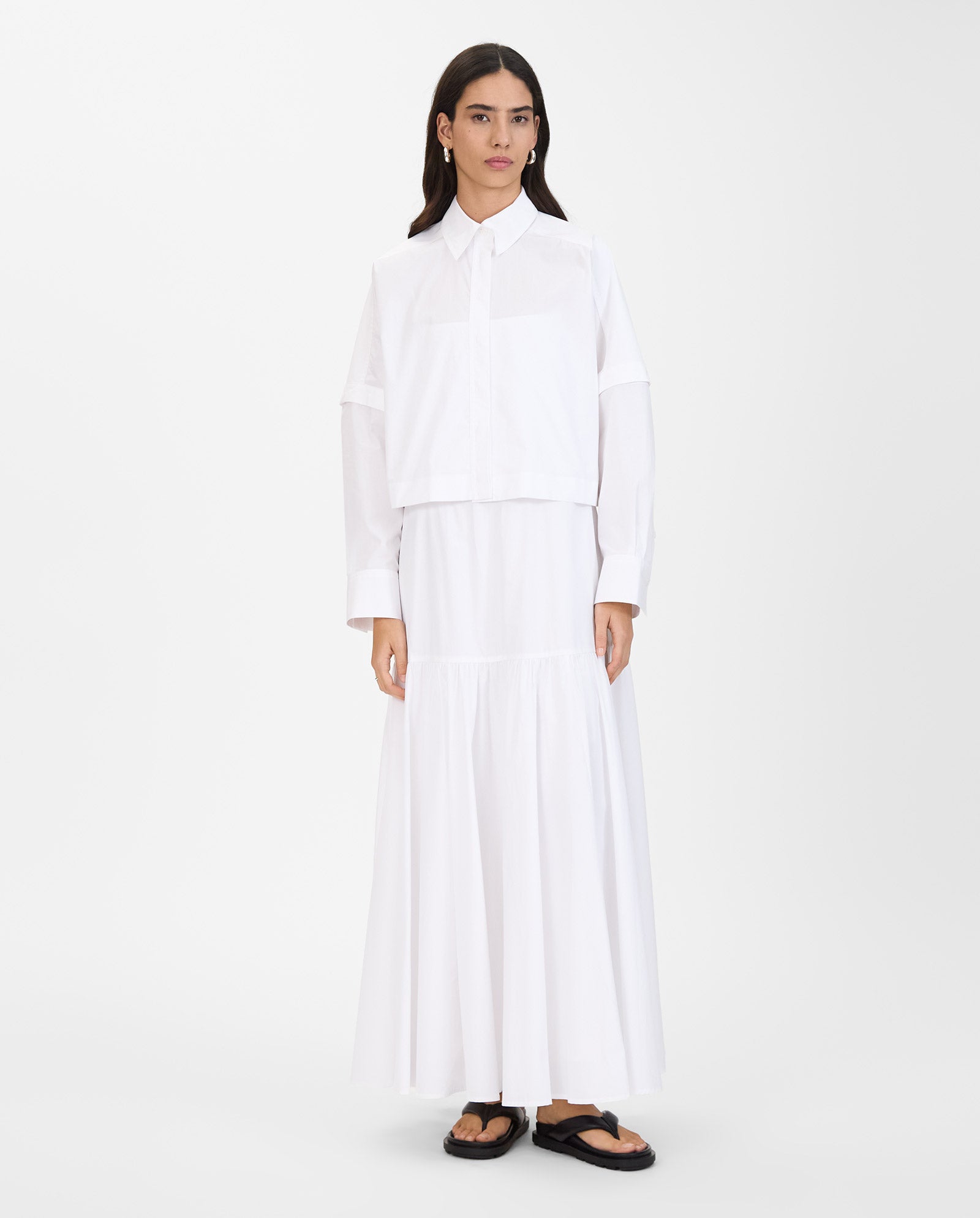 Woman stands in a long white dress, IVY OAKs ELVIRA Shirt with detachable sleeves, and black sandals against a plain white background.